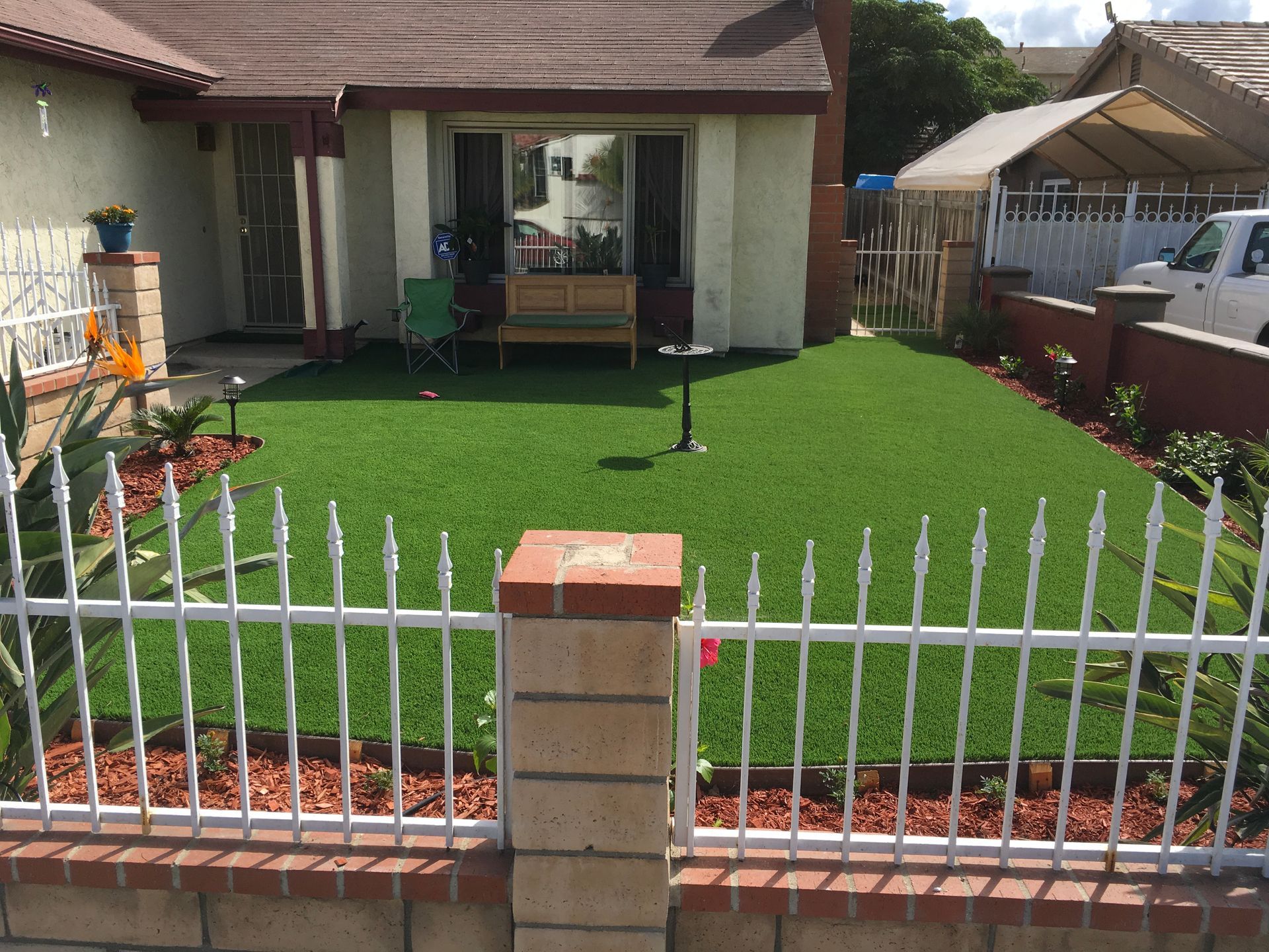 A house with white fence, brick details, and green artificial turf lawn. Red mulch borders the yard.
