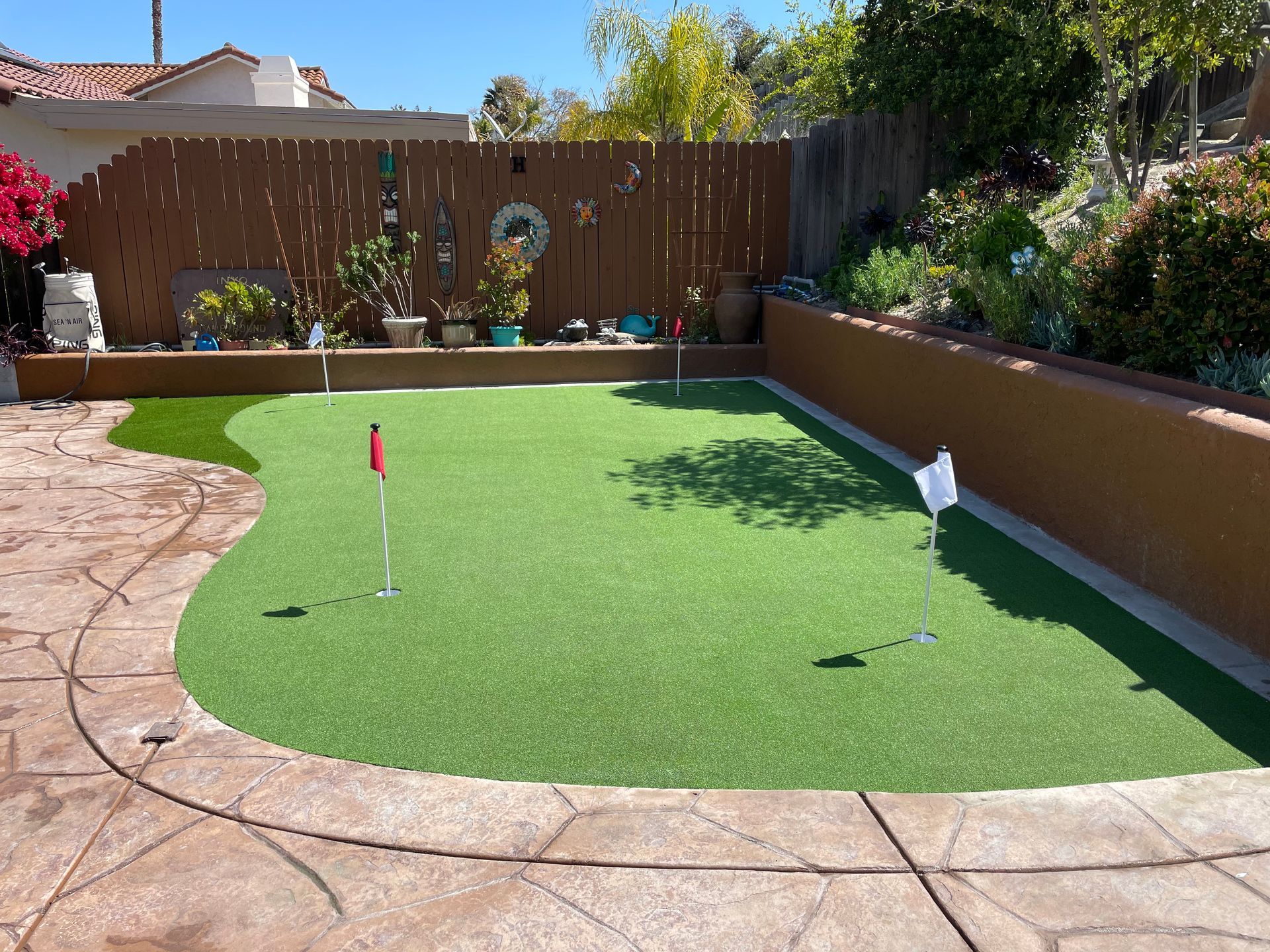 Backyard putting green with three holes and flags, surrounded by stone patio and a brown fence.