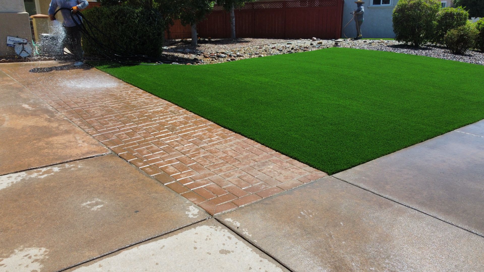A person spraying water on a brick and concrete driveway next to green artificial turf.