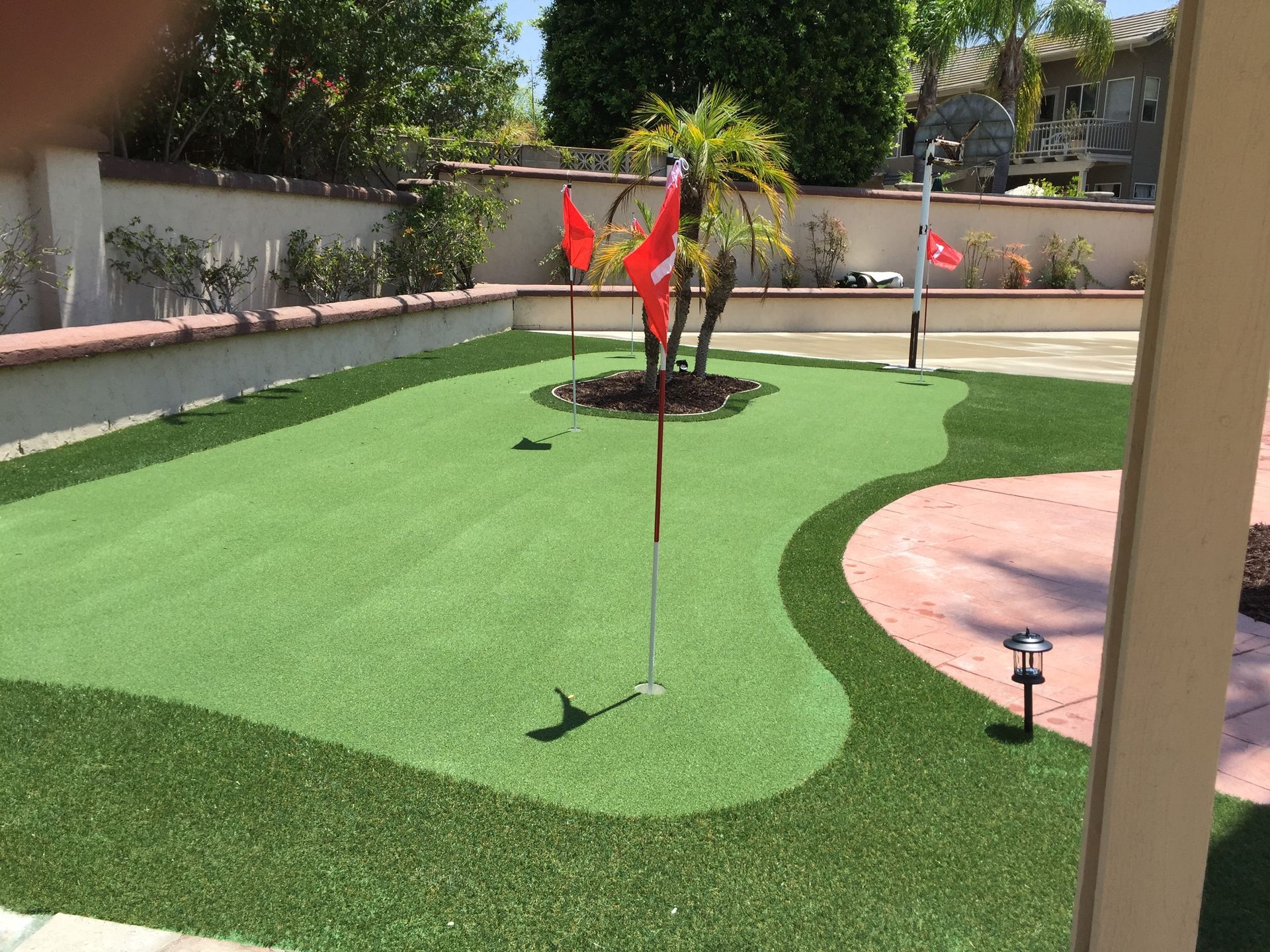 Backyard putting green with three holes and red flags. Artificial turf and red brick patio.