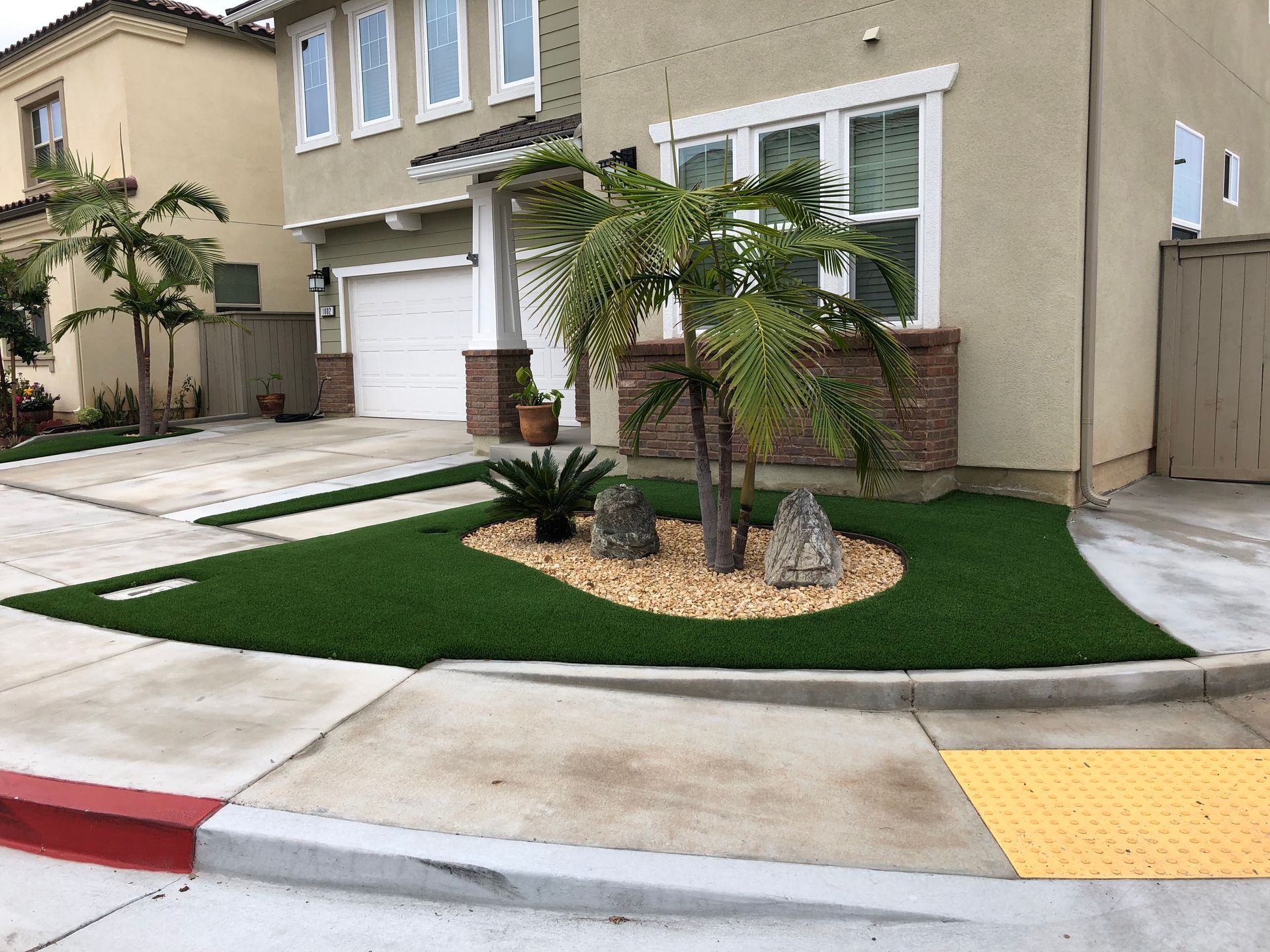 A two-story house with green artificial turf landscaping, a palm tree, and a concrete driveway.