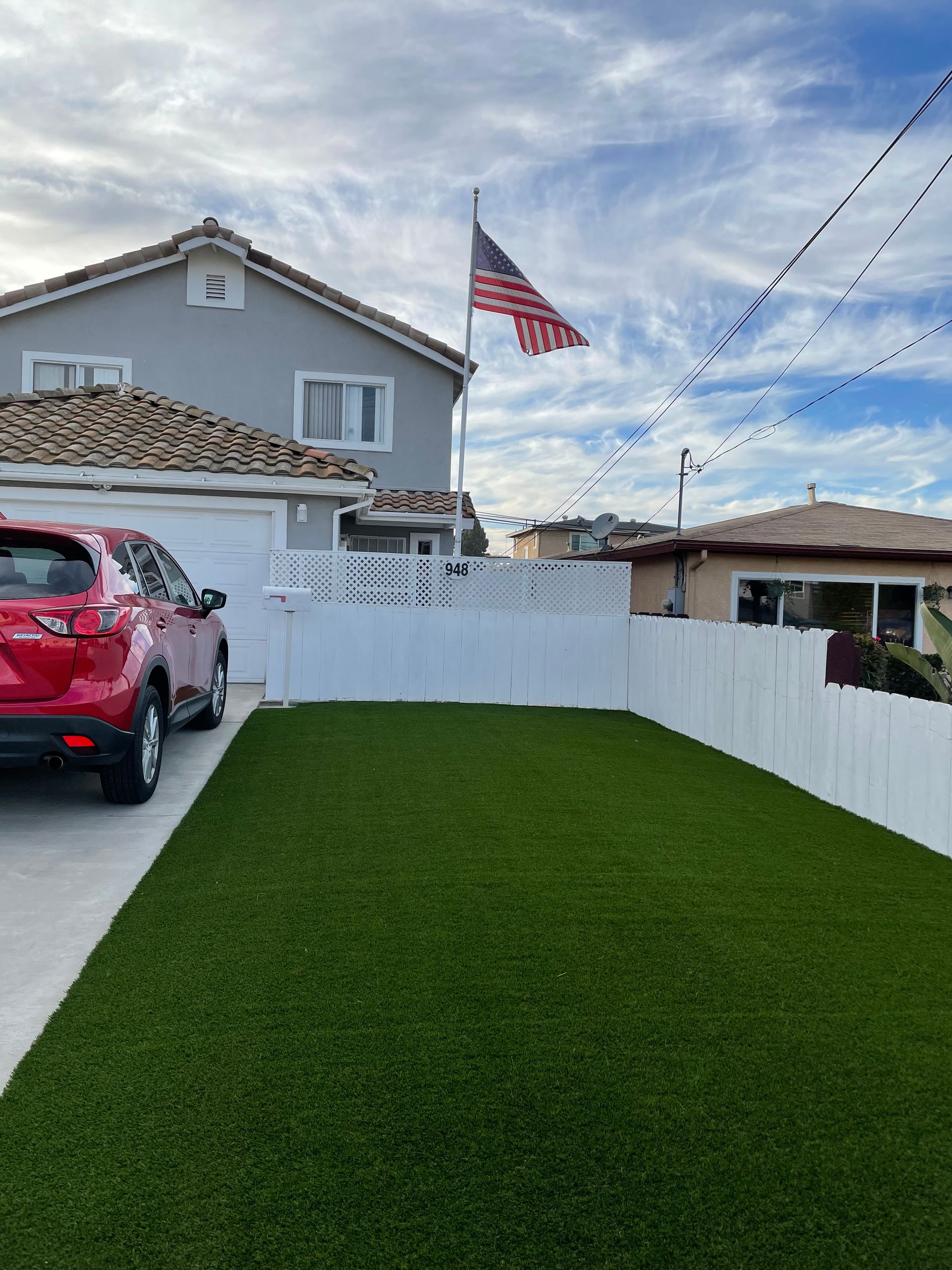 Red car parked in front of a house with American flag flying, white fence and green lawn.