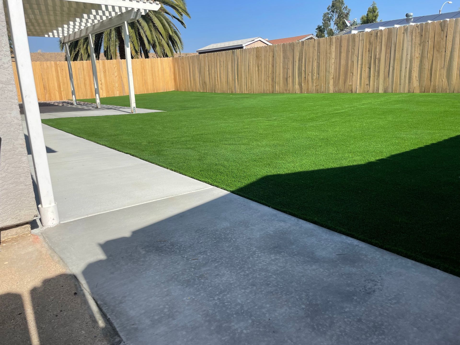 Concrete patio leads to a backyard with green turf, wooden fence, and a pergola. Sunny day.