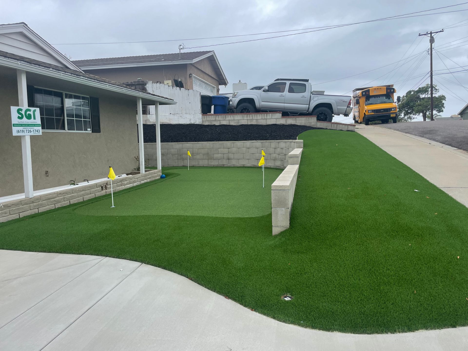 Green artificial turf putting green in front of a house, with golf flags and a truck parked above.