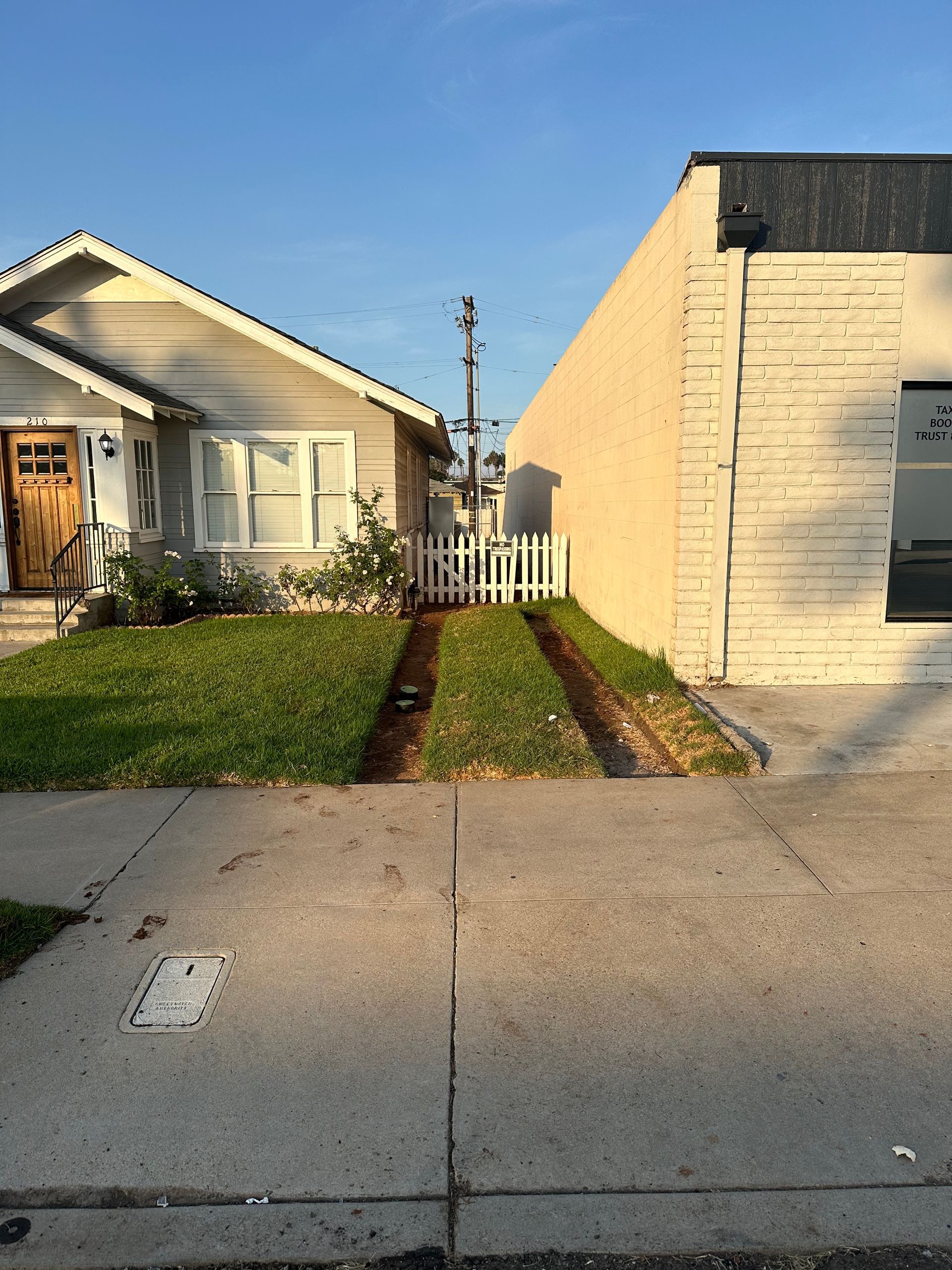 A sidewalk leads to a narrow driveway between a house and building, with tire tracks in the grass.