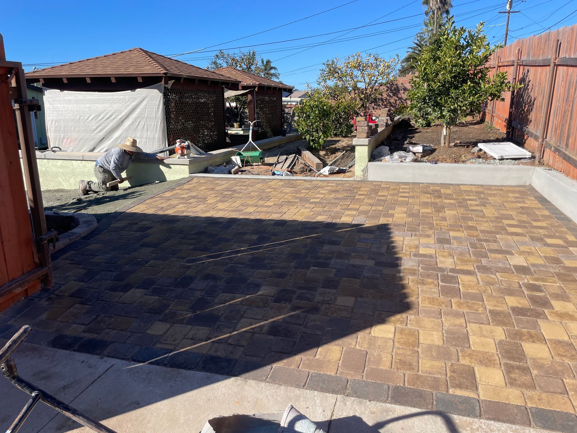 Brick patio being constructed in a backyard, person working, brown and tan pavers, brown fence, clear sky.