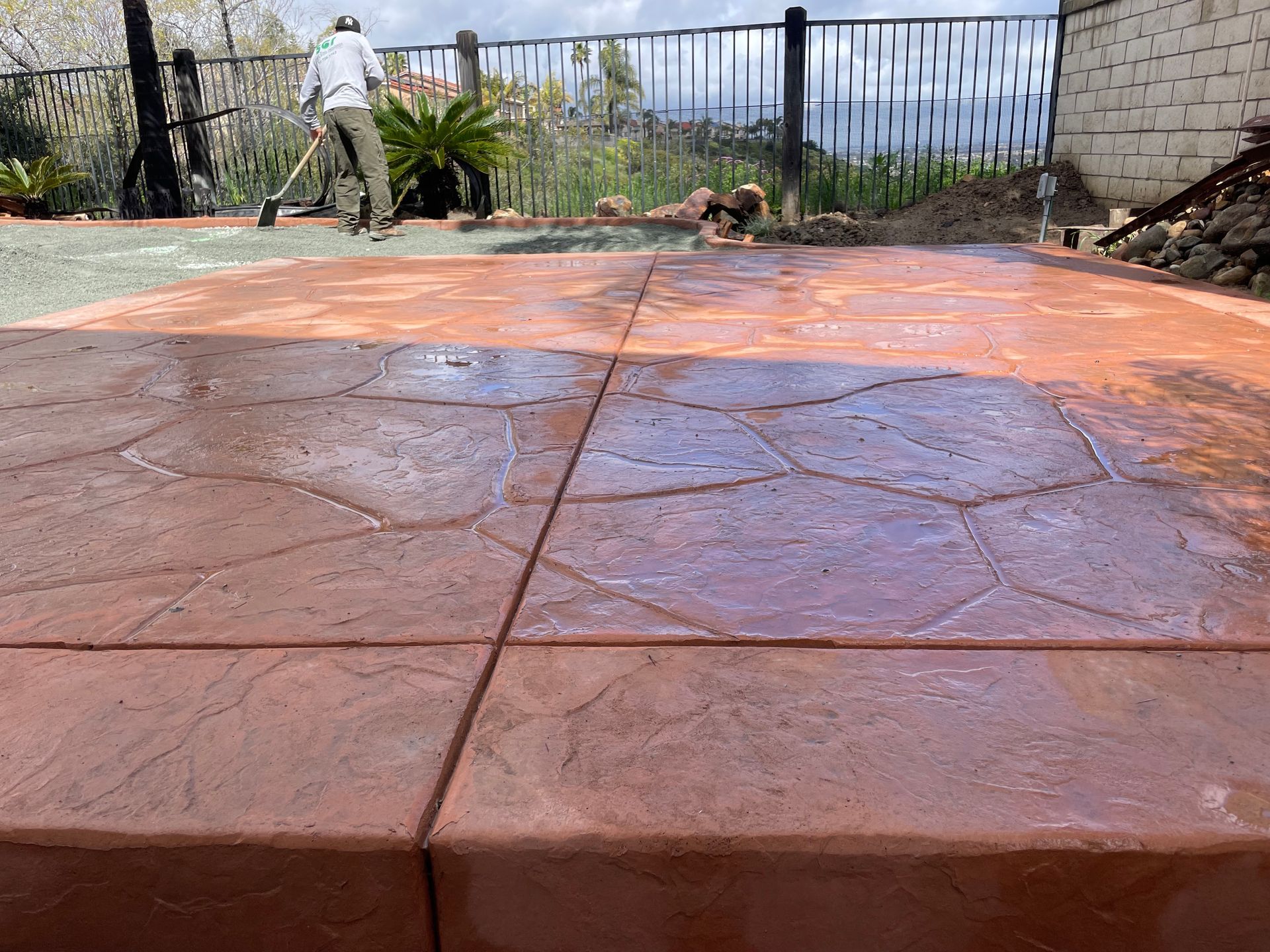 Man applying sealant to a wet, reddish-brown stone-patterned concrete patio. Fenced yard in background.