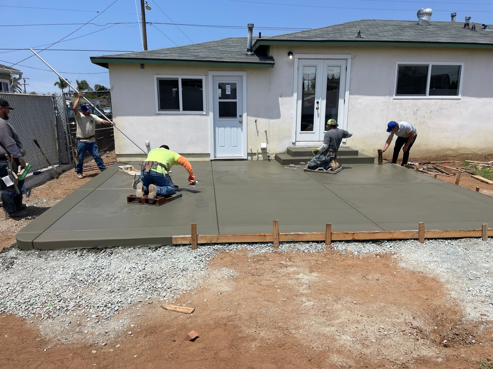 Construction workers pouring and smoothing concrete for a patio outside a house on a sunny day.