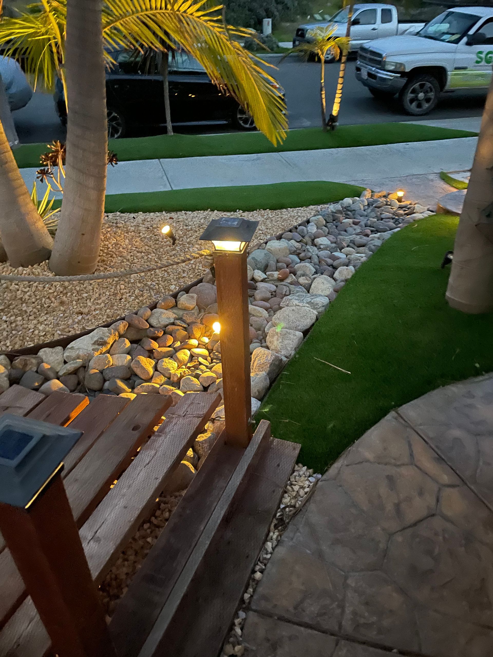Outdoor scene with illuminated pathway, stairs, and landscaping including palm trees, rocks and artificial grass.