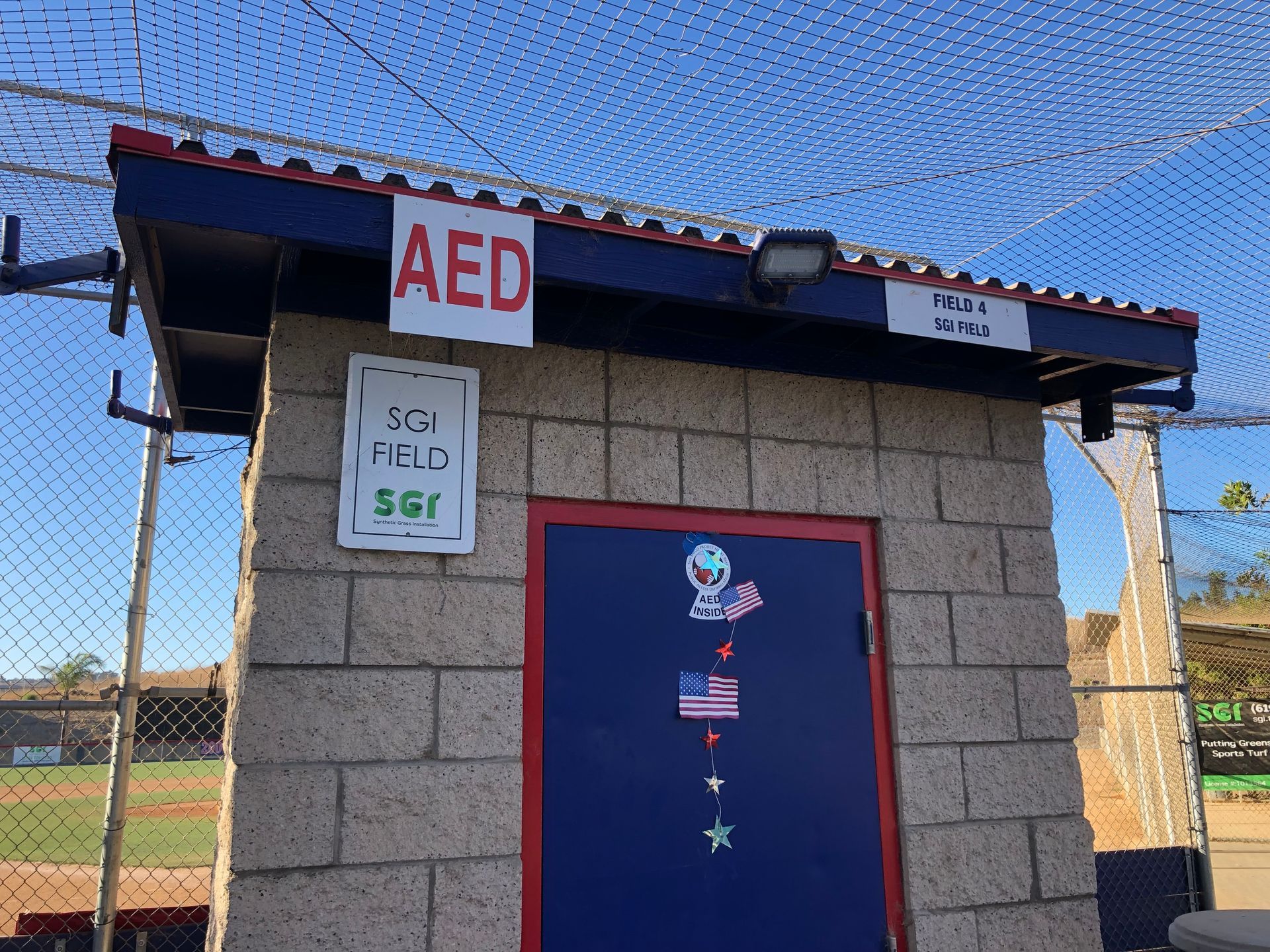 Small brick building with red door, AED sign, and baseball field in the background.