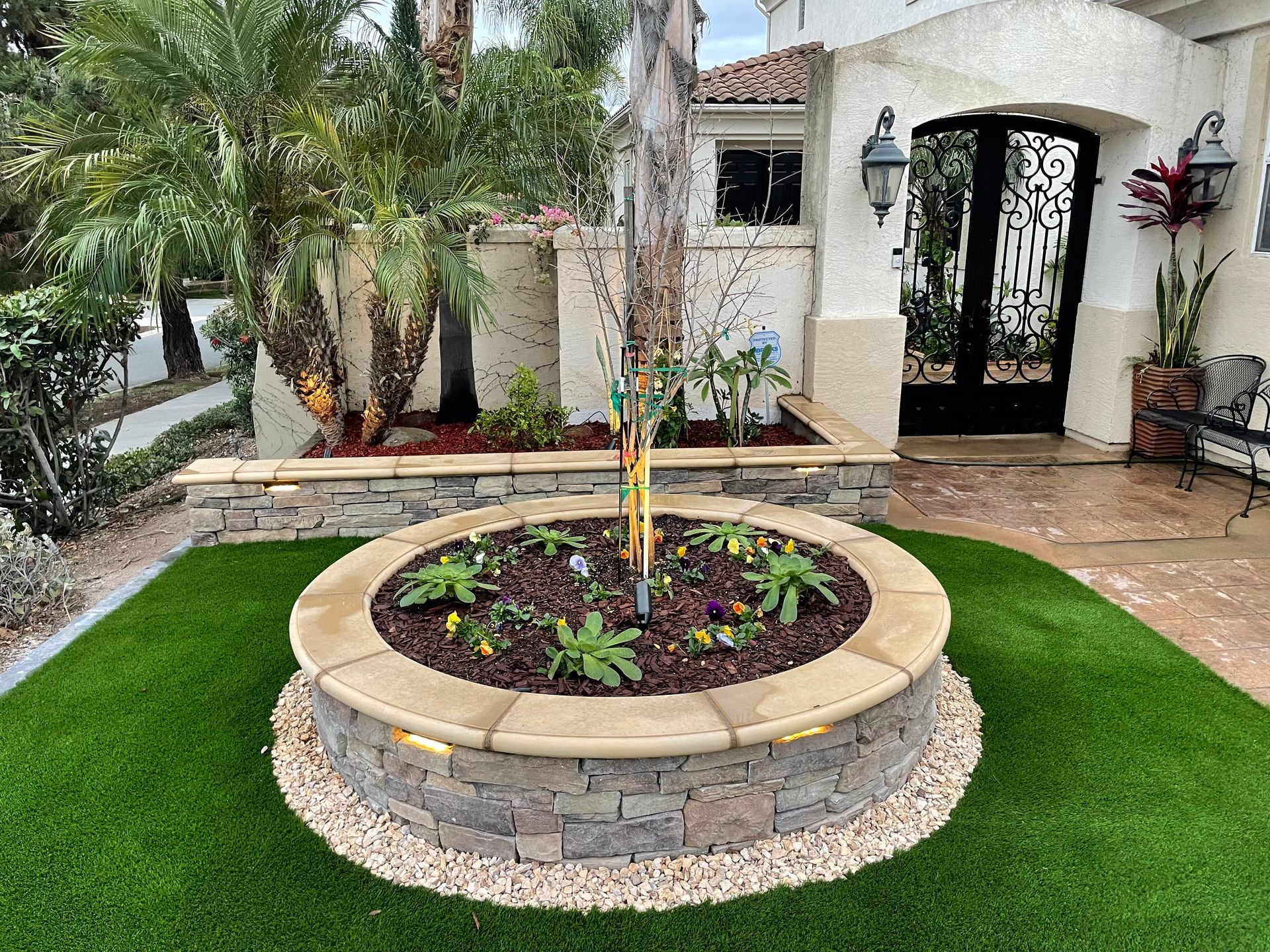 Circular stone planter with small tree, surrounded by grass and pebbles, in front of a house.