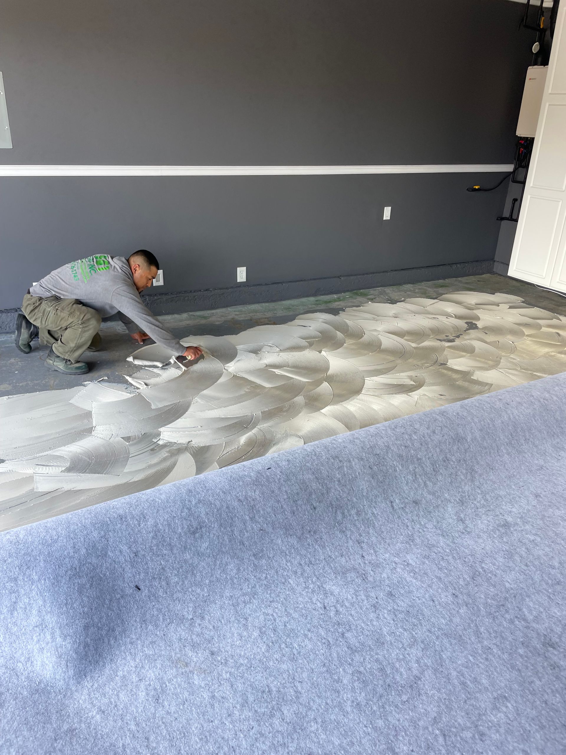 A person applying a coating to a concrete garage floor. Gray walls, white trim, and a blue floor covering.