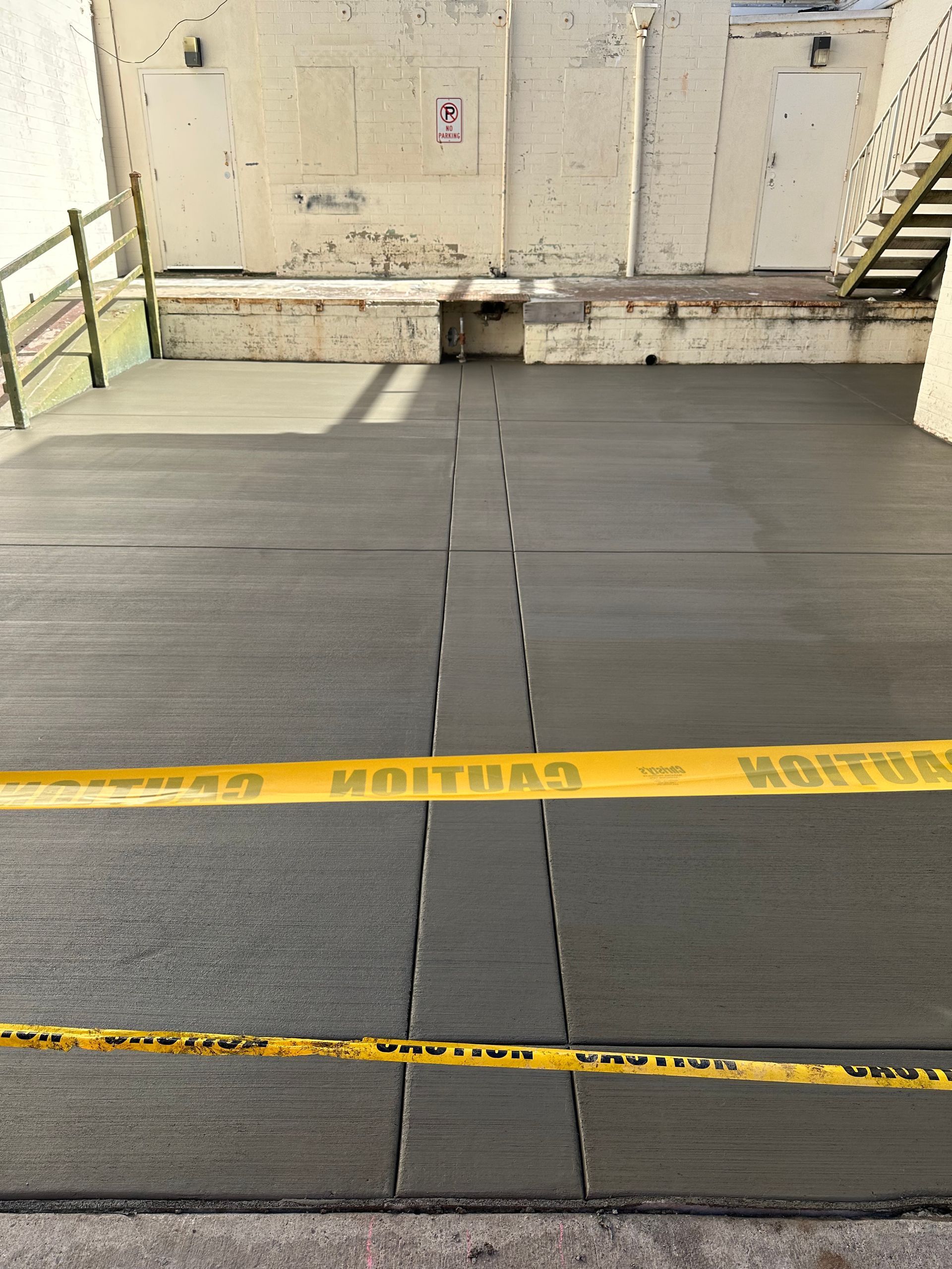 Freshly poured concrete patio with yellow caution tape, against a light-colored building with stairs.