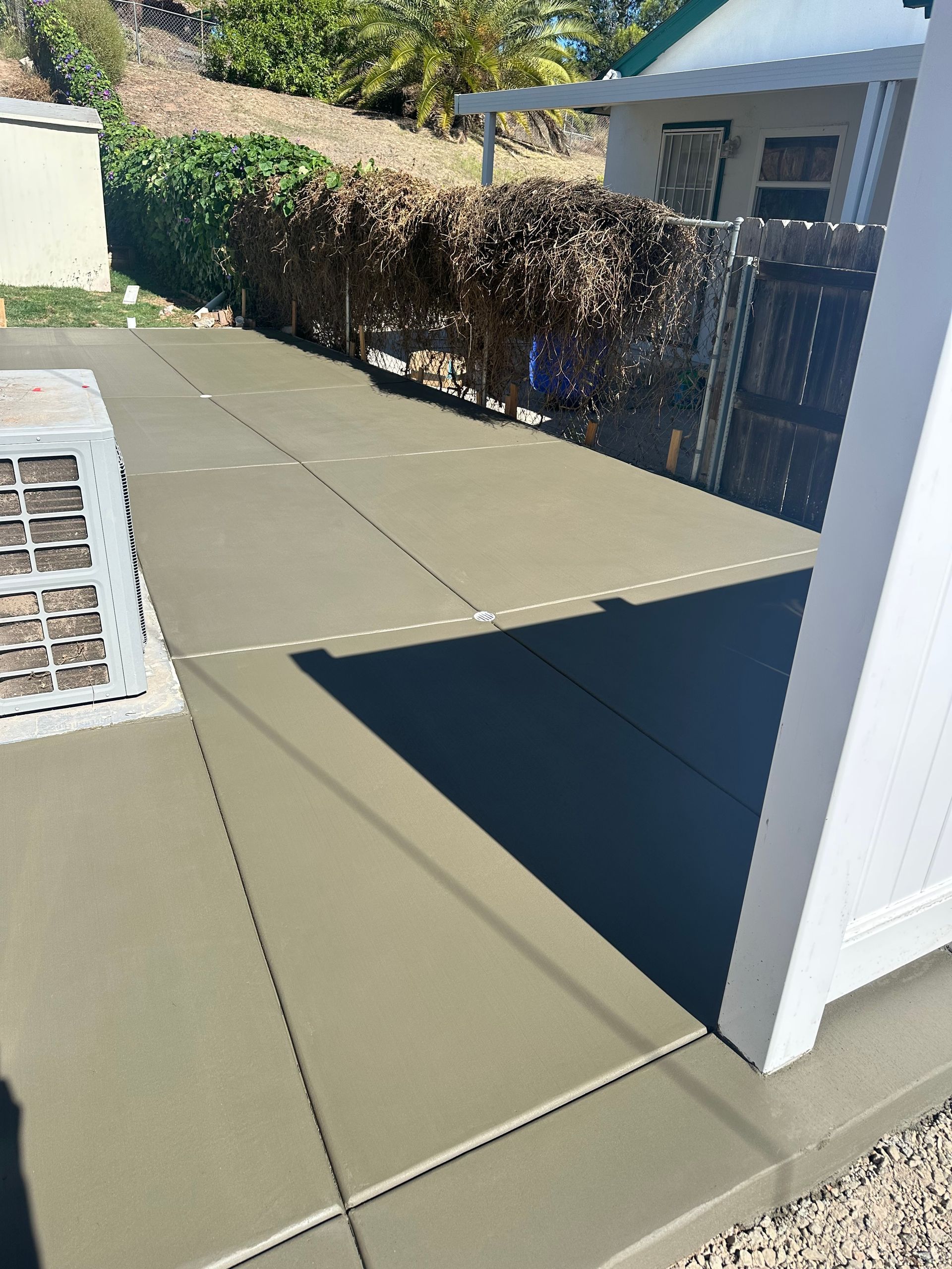 Newly poured concrete patio next to a white fence. Green vegetation and small building in the background.