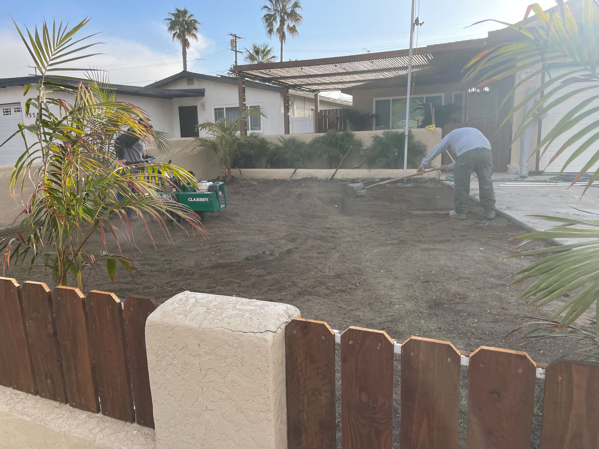 Man spreading soil in a yard in front of a house, wooden fence in foreground, palm trees in the background.
