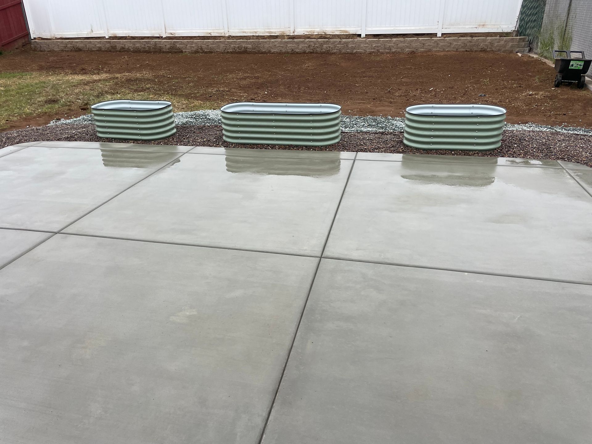 Three green metal planters sit on a concrete patio, with a gravel border and a dirt area behind.