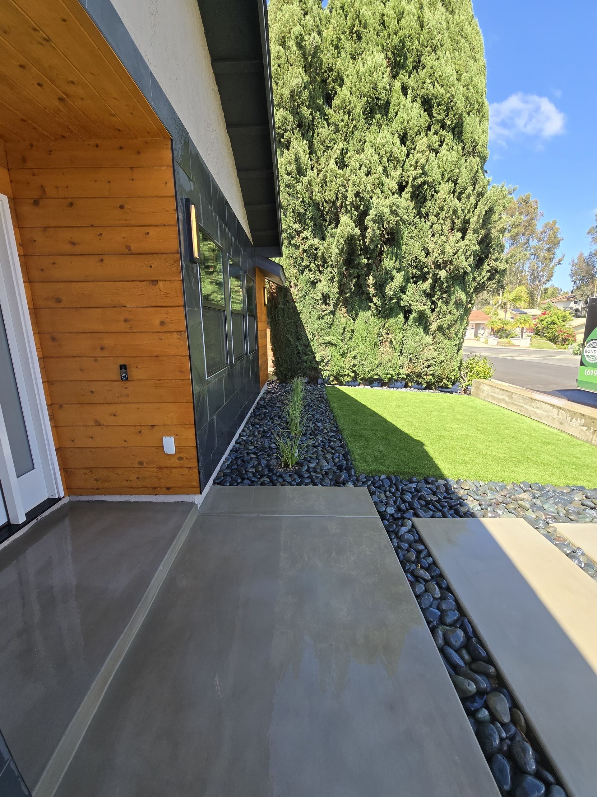 Exterior view of a modern house with a concrete walkway, black rock garden, and wood siding.