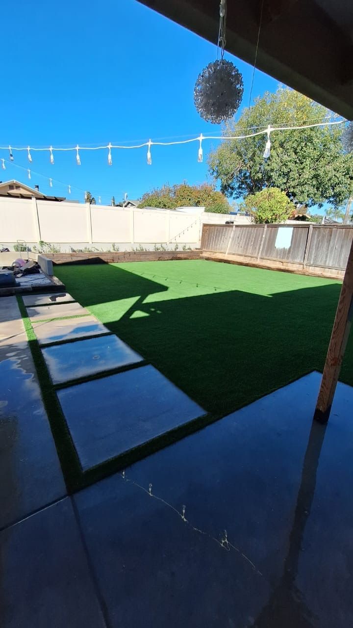 Backyard with green artificial grass, concrete patio, stepping stones, and string lights against a blue sky.