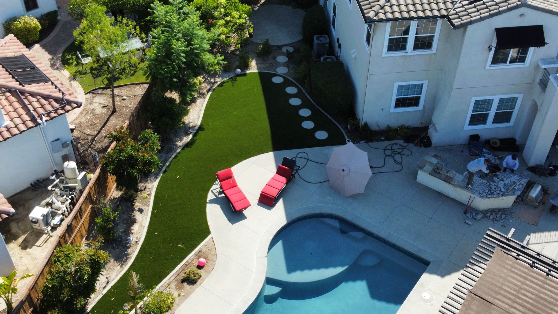 Aerial view of a backyard with a pool, lounge chairs, patio, and artificial grass. A two-story house is nearby.