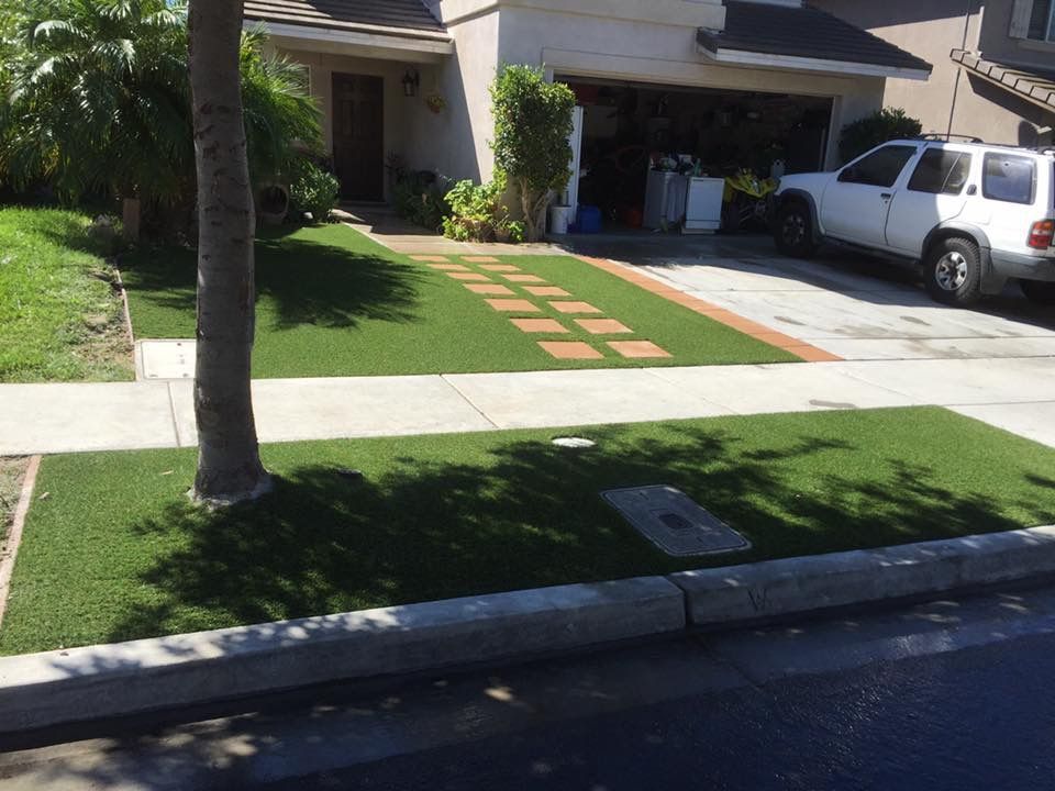 A home's artificial turf yard with tree, driveway with stepping stones, and a white SUV parked in the garage.