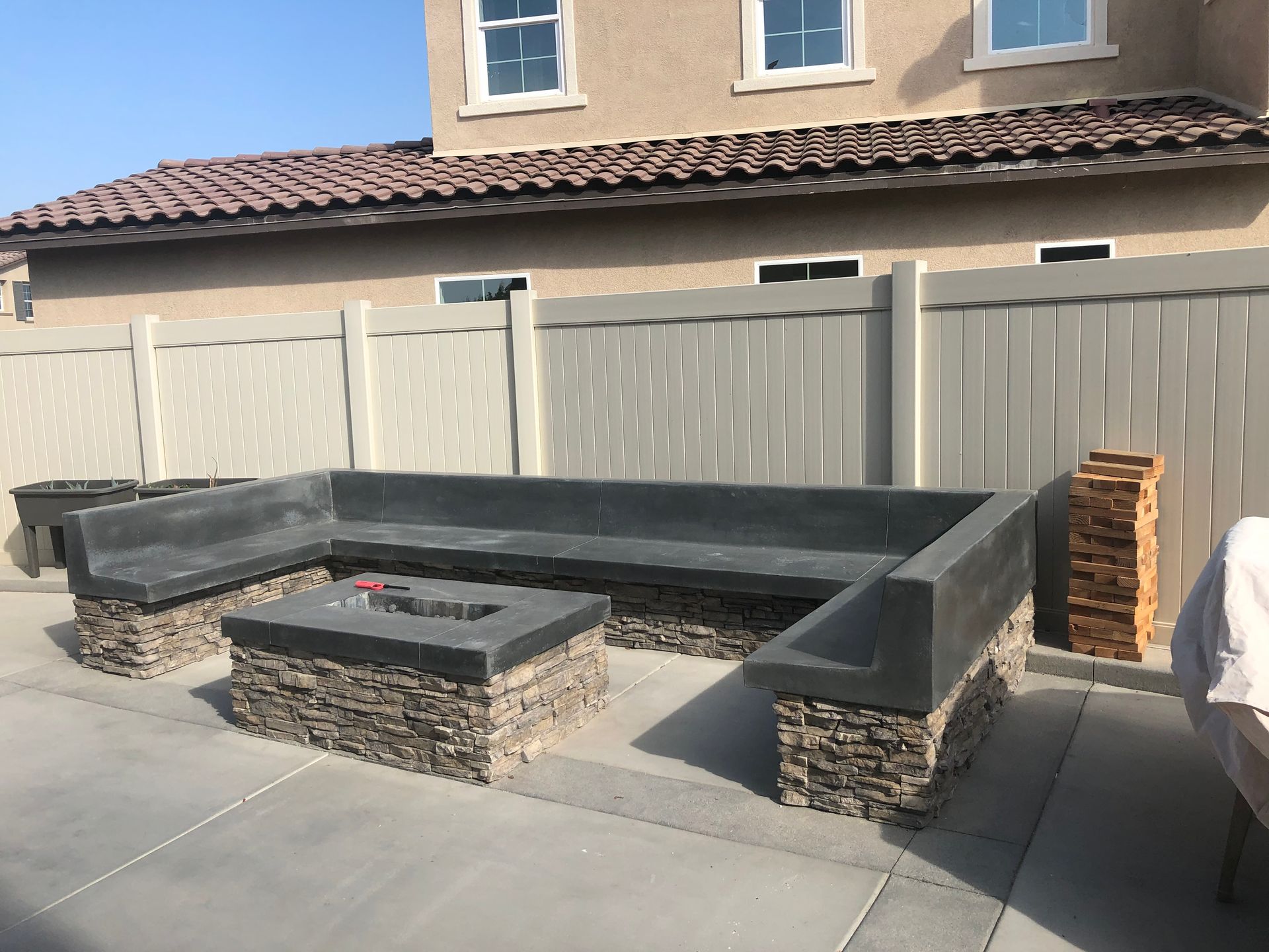 Patio with stone and concrete seating around a fire pit, next to a beige fence and house.