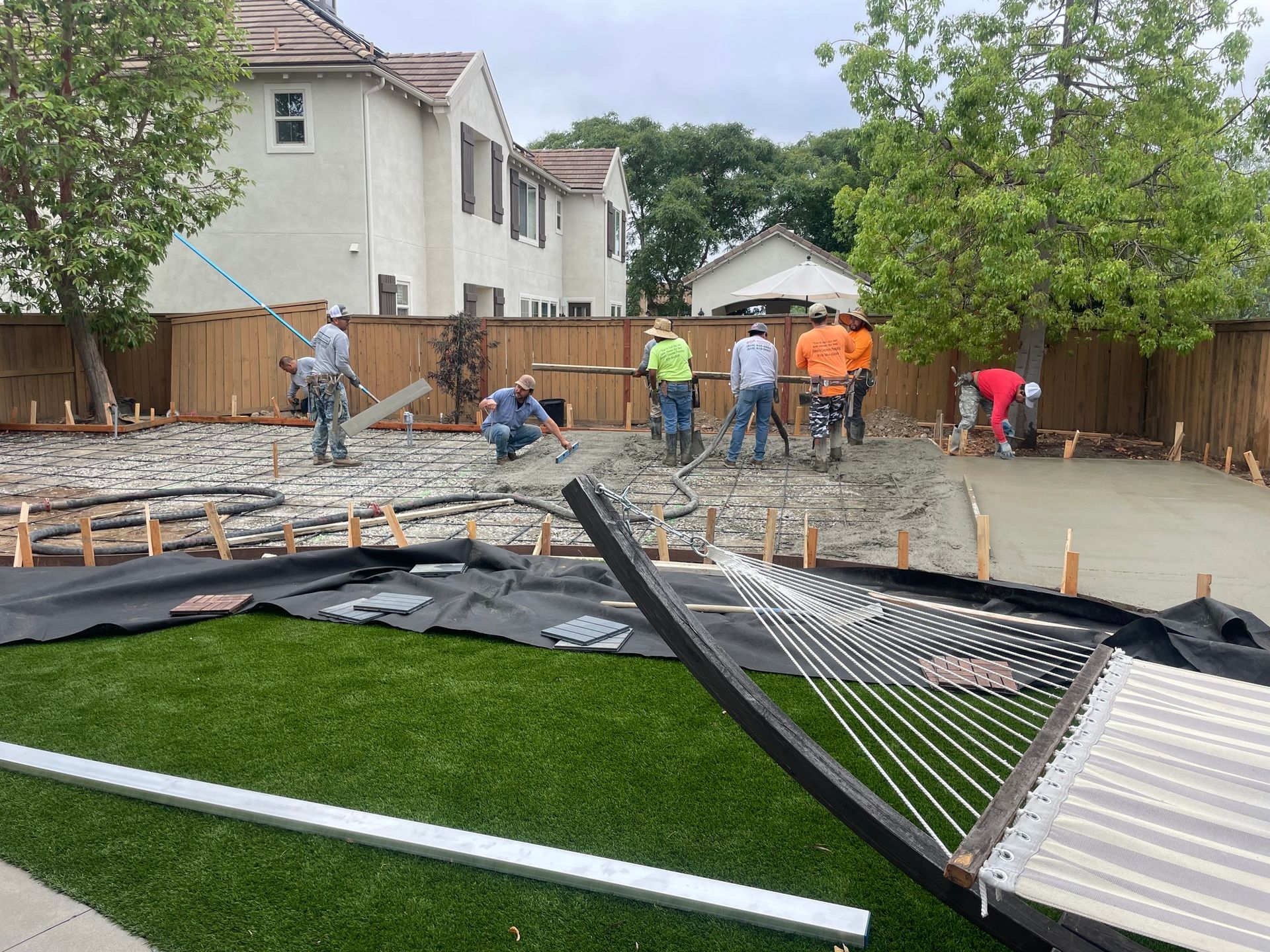 Construction workers pouring concrete in a backyard, surrounded by a wooden fence, with green grass in foreground.