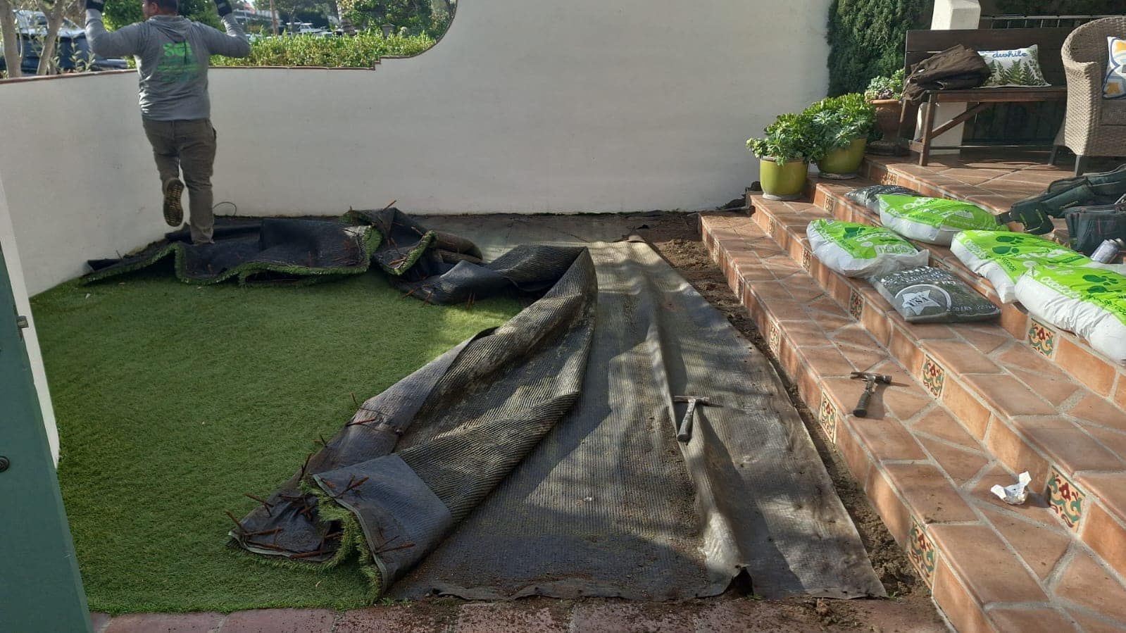 Man laying artificial turf in an outdoor courtyard with brick steps and patio furniture.