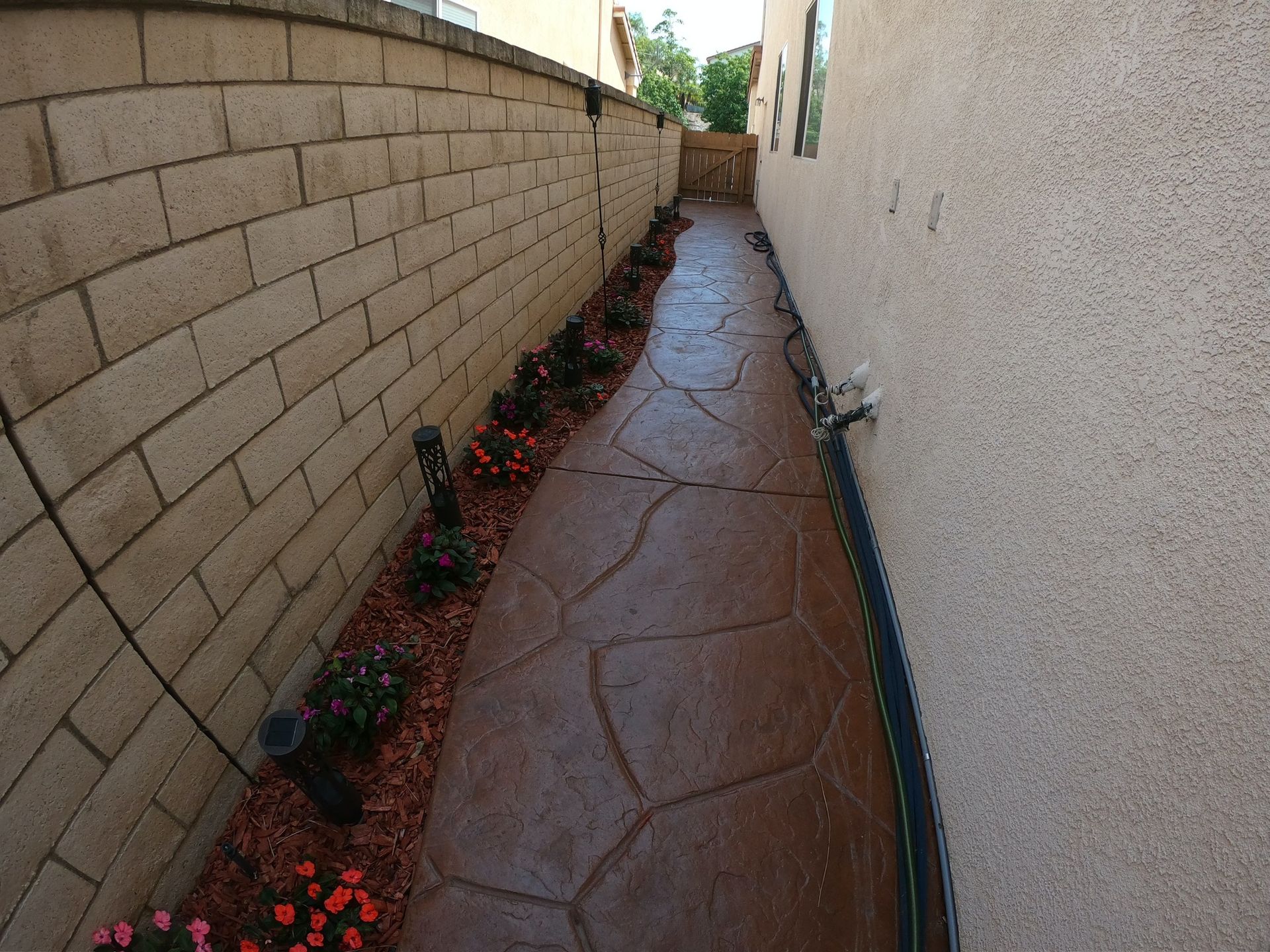 Narrow walkway with brick wall and textured brown concrete path lined with flowers.