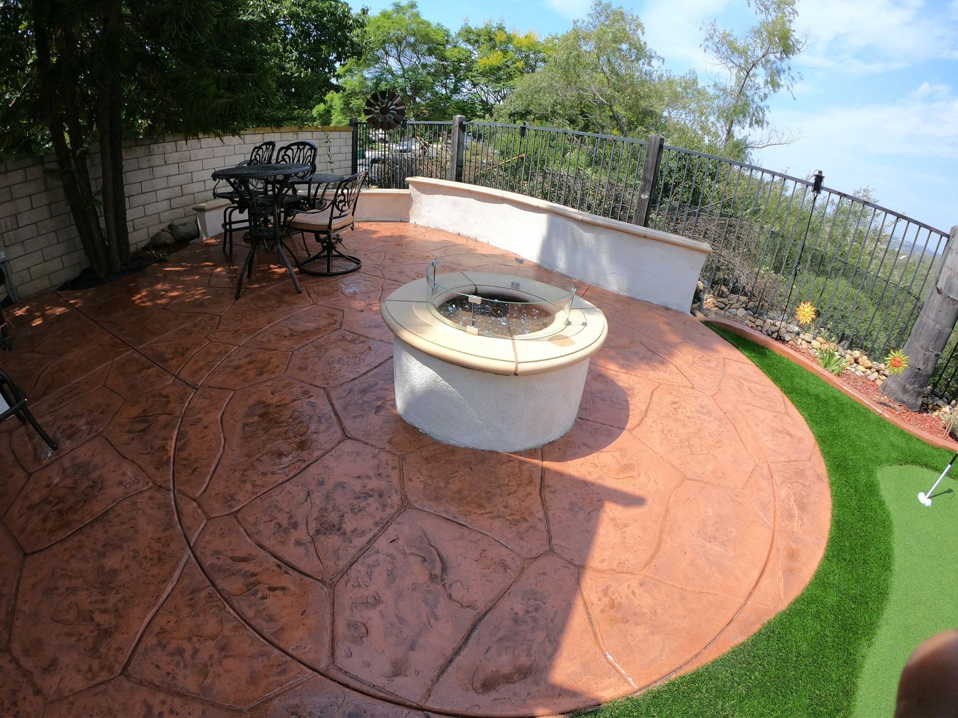 Patio with concrete fire pit, dining set, and putting green. Brown stamped concrete. Sunny outdoor setting.