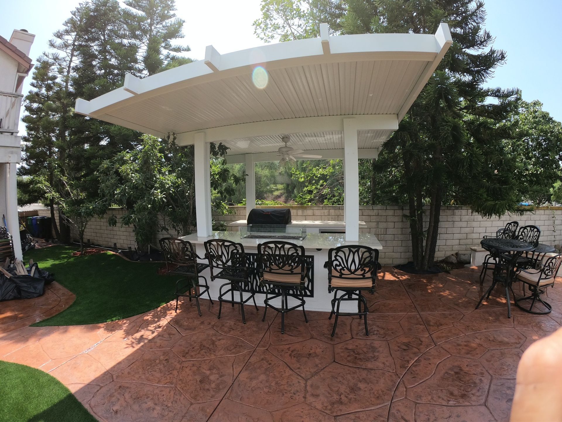 Outdoor kitchen with overhead structure, built-in grill, bar seating, on stamped concrete patio.