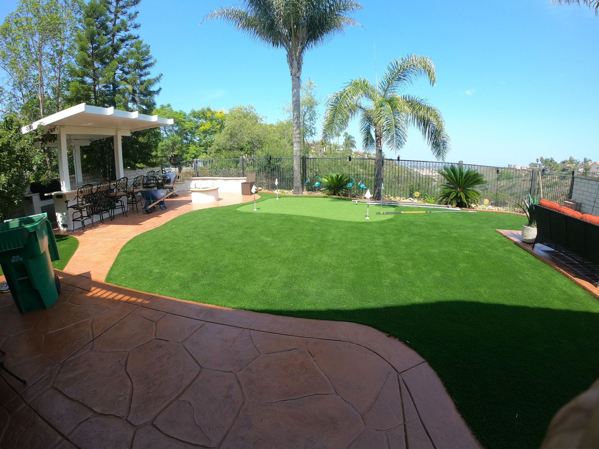 Backyard with green turf, putting green, palm trees, and a patio with a covered grilling area.