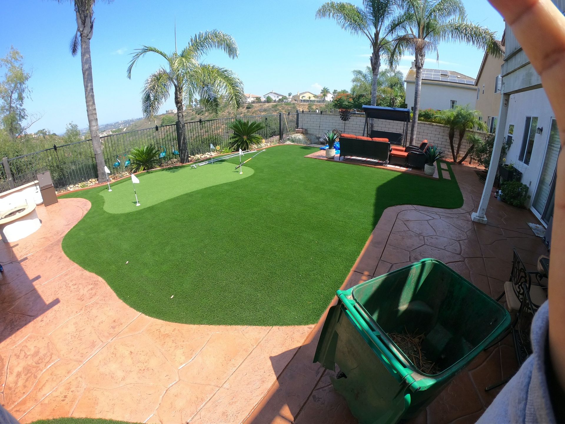 Backyard with putting green, palm trees, and a green trash bin on a stamped concrete patio.