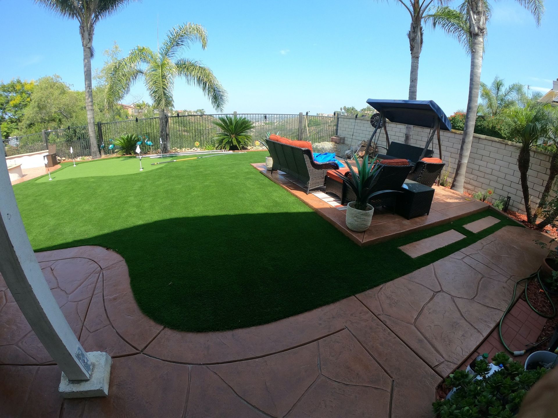 Backyard with artificial turf, patio seating, and palm trees under a blue sky.