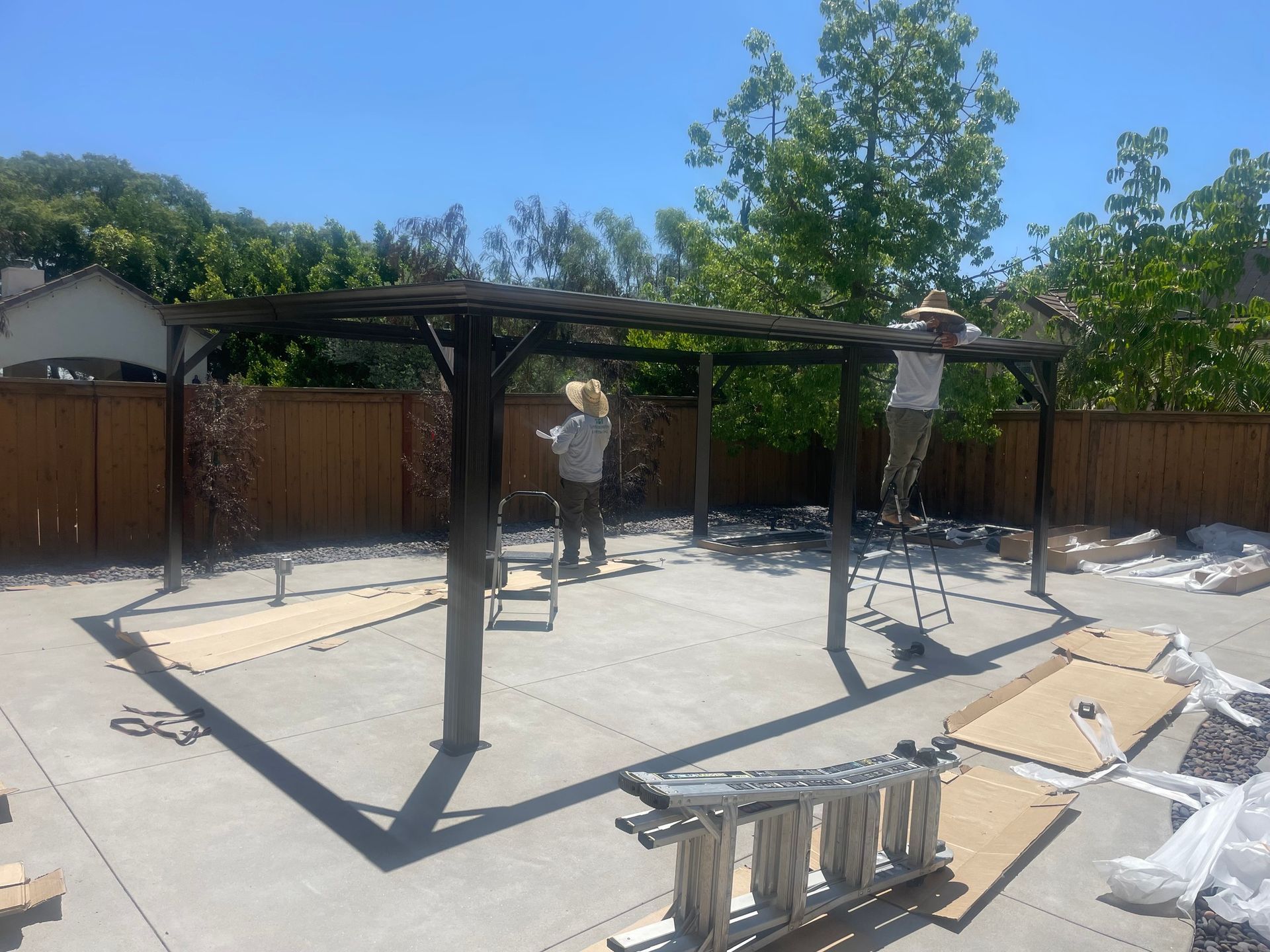 People assembling a dark brown pergola on a concrete patio, with a wooden fence and trees in the background.