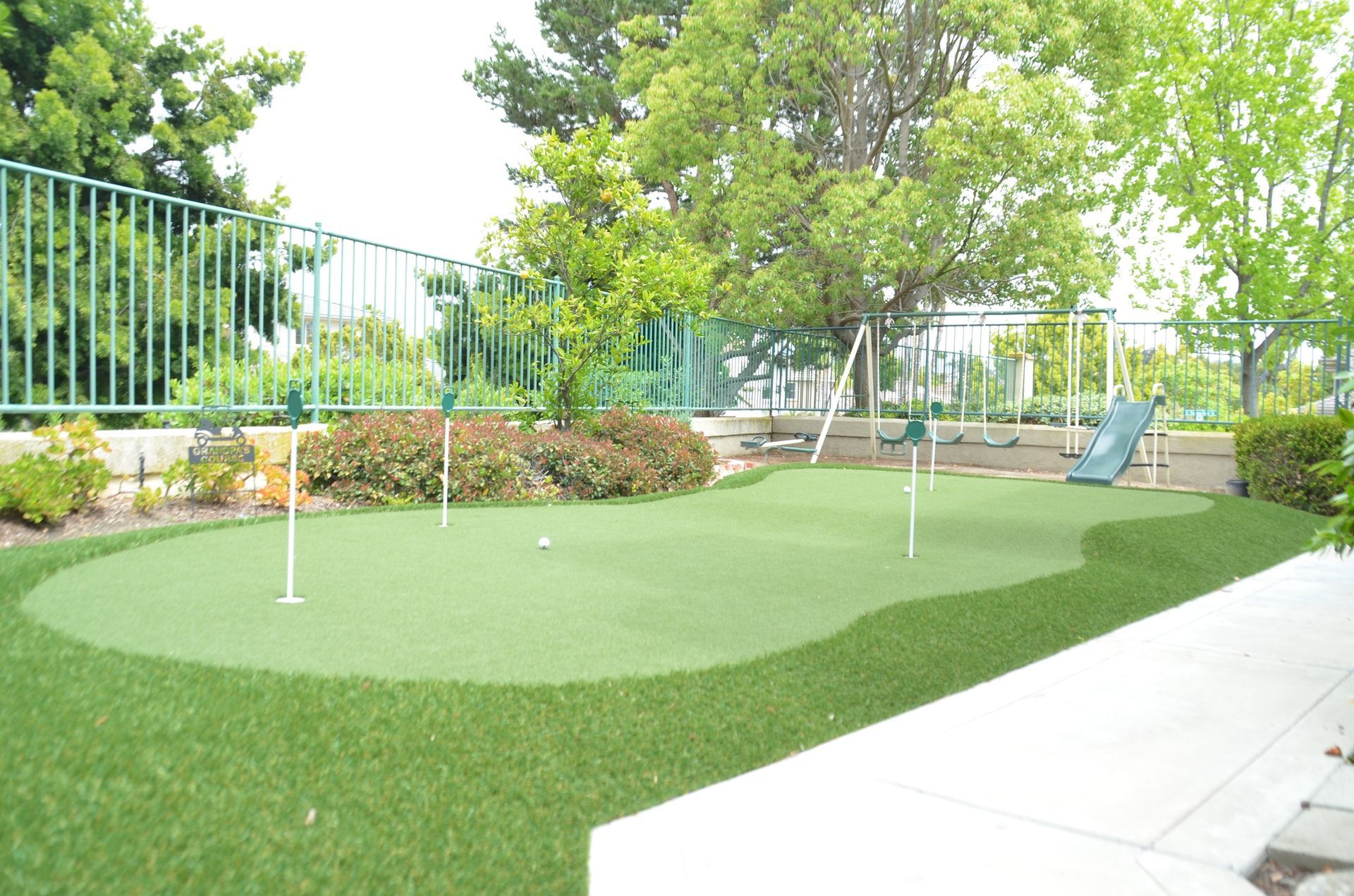 Green putting green with two flags, overlooking a playground, fence, and trees.