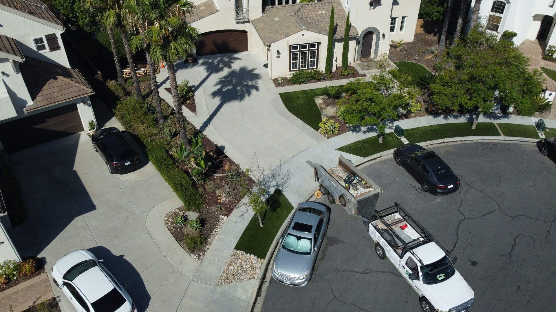 Aerial view of several cars parked on a residential street. A large house with a circular driveway is in the background.