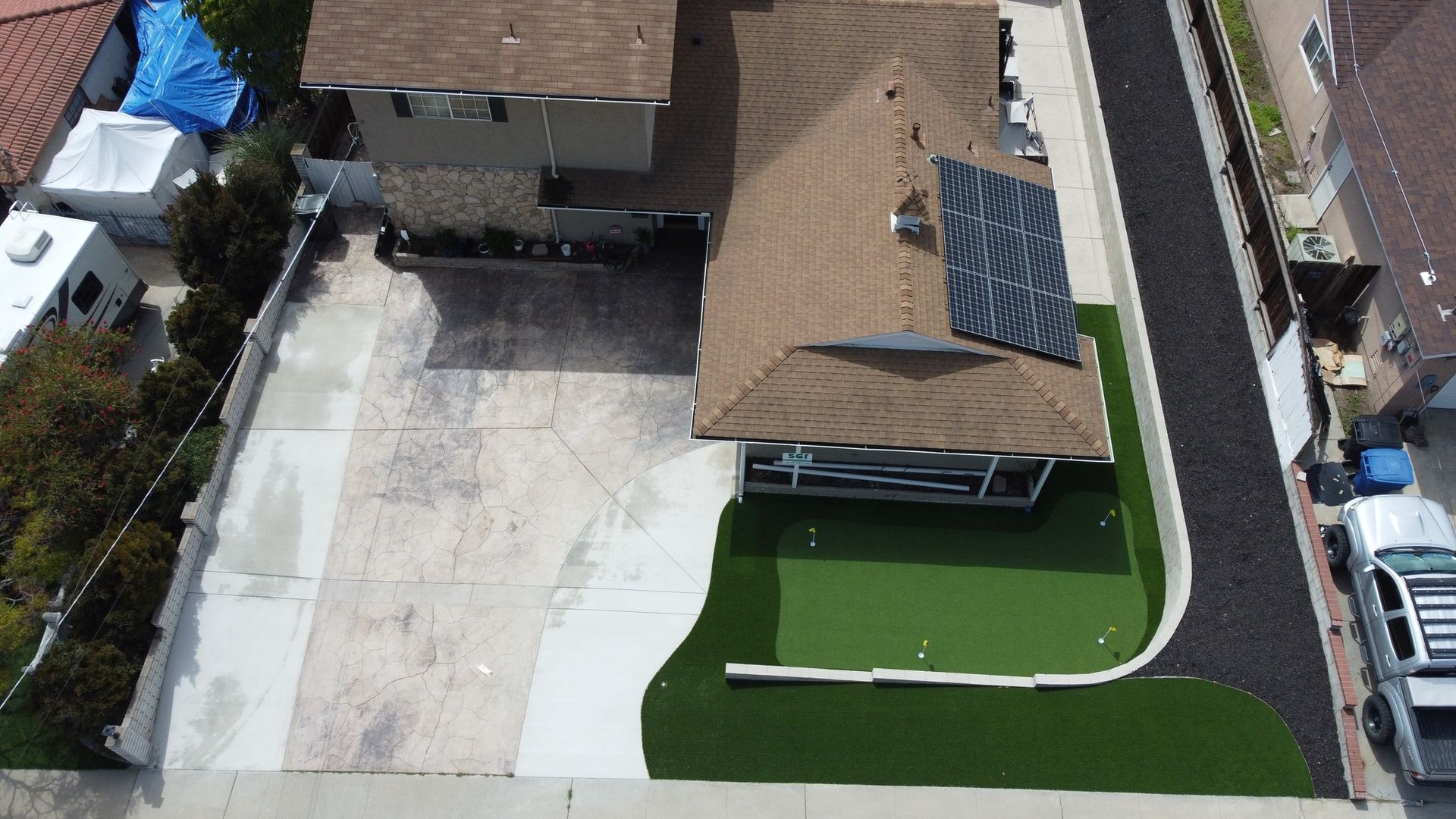 Aerial view of a house with a brown roof, solar panels, and a concrete driveway. Green lawn borders the drive.