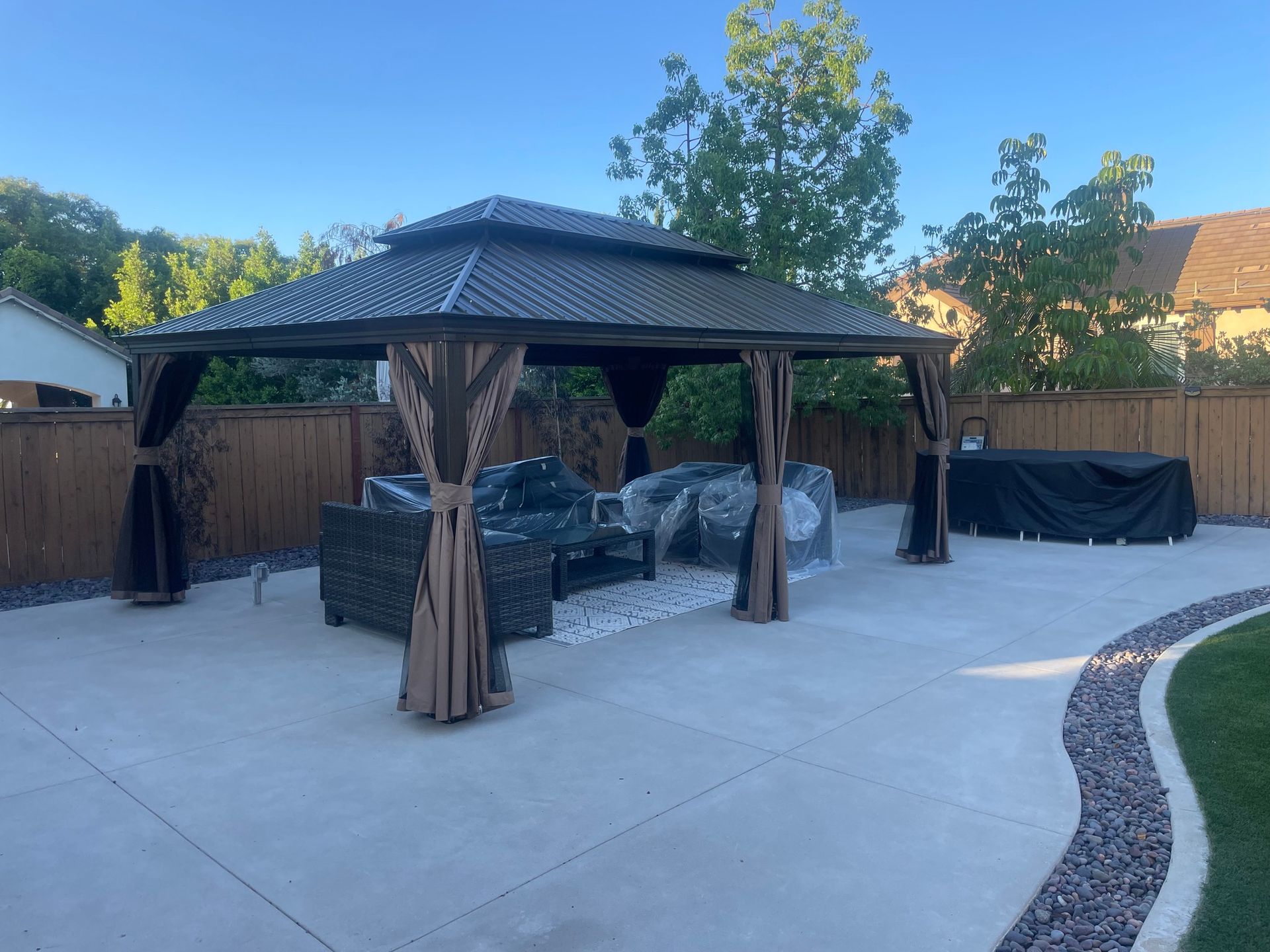 Gazebo with brown roof, curtains, and patio furniture on a concrete patio, with a wooden fence background.
