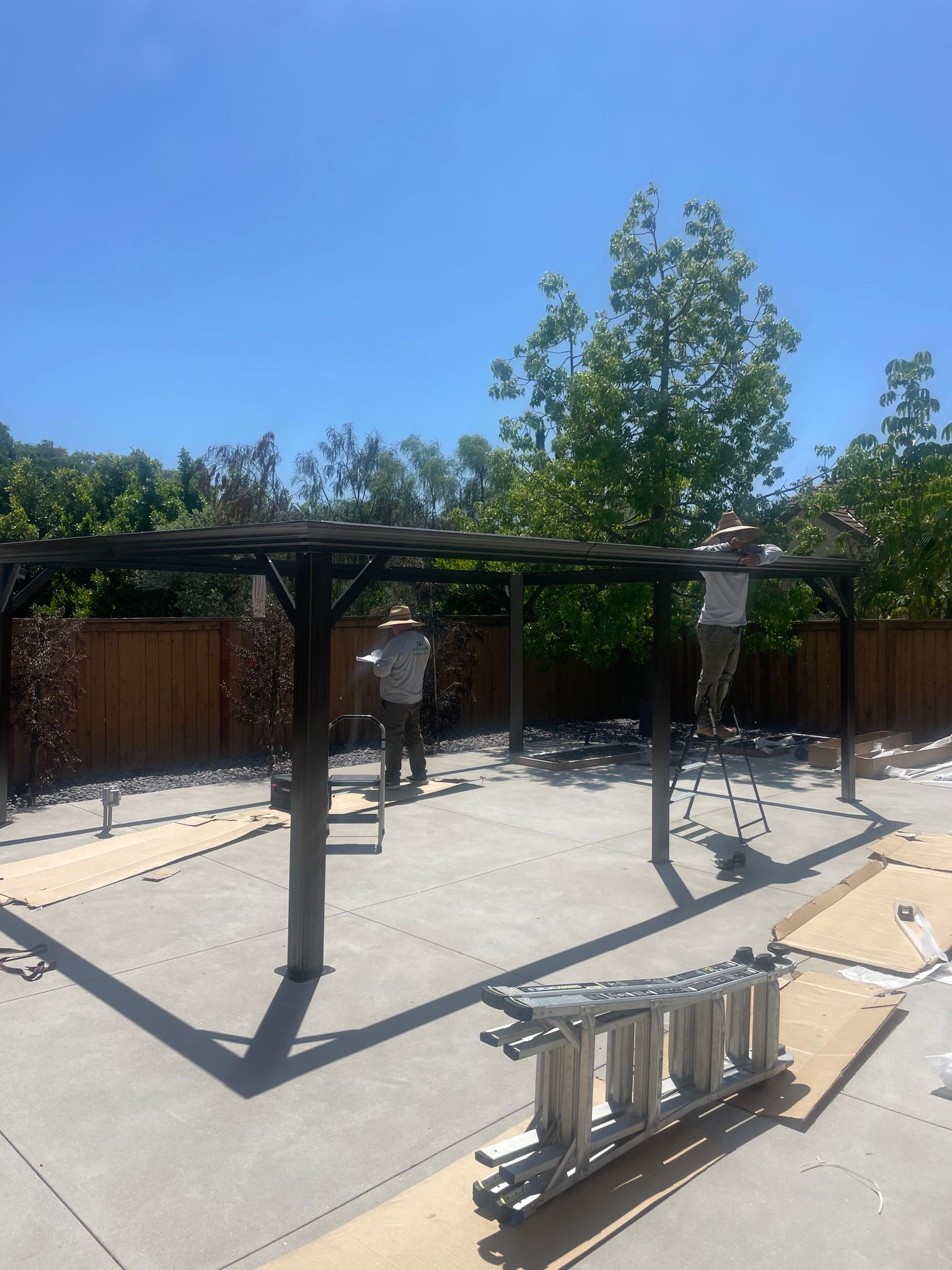 People constructing a dark-stained wooden pergola in a backyard on a sunny day.