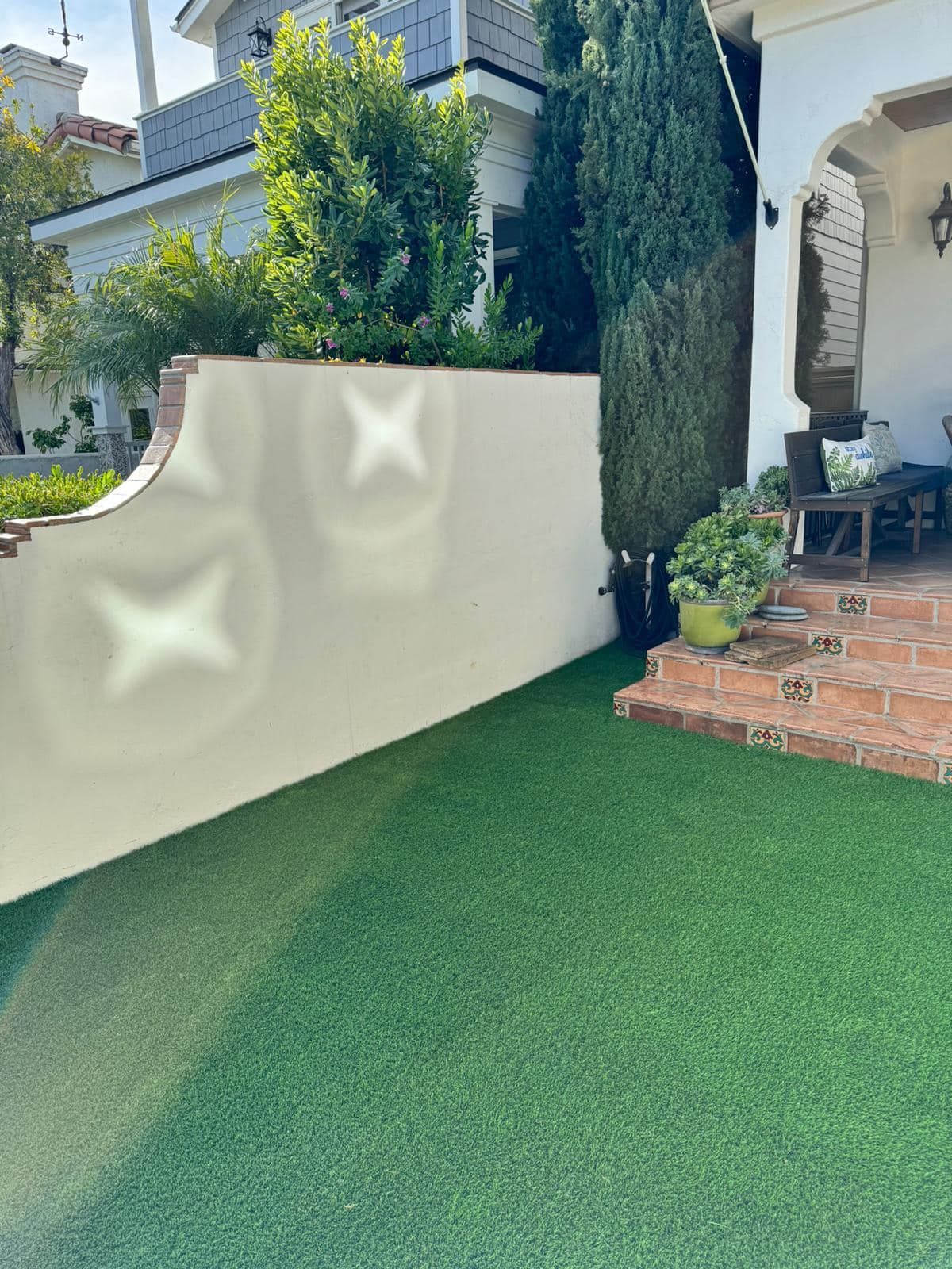 A sunny front yard with green turf, a white stucco wall, and a house with red brick steps.
