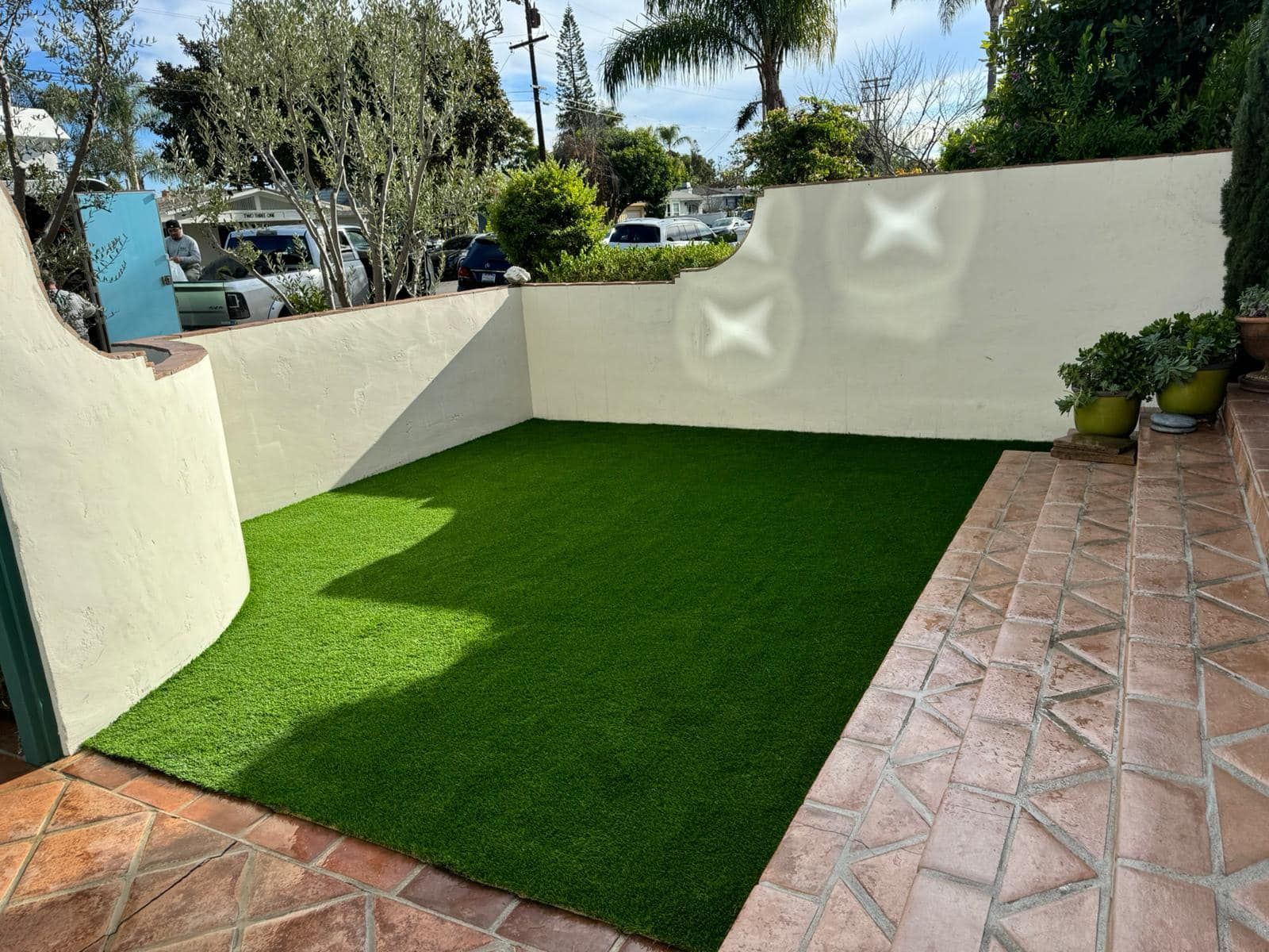 Artificial turf in a walled yard with brick pathway and potted plants.