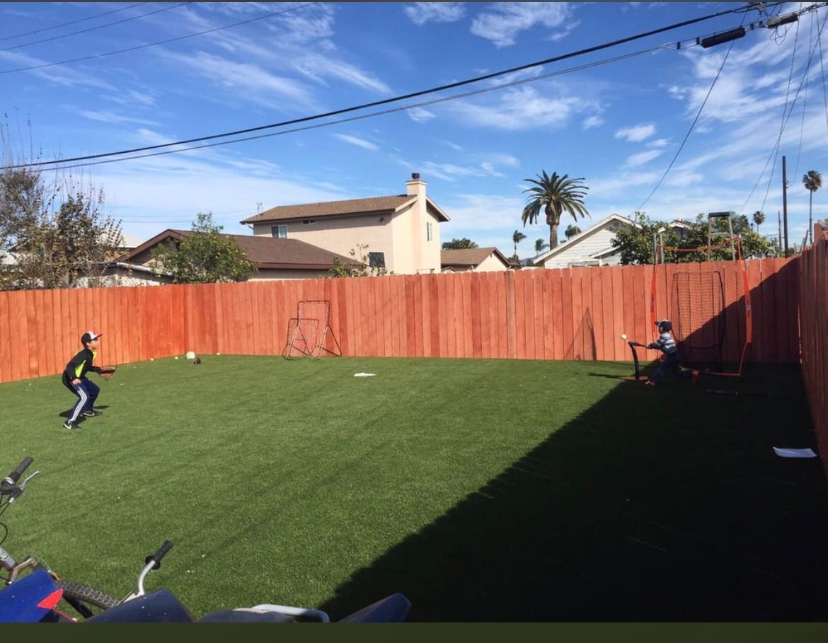 Two people play baseball in a fenced backyard with artificial turf on a sunny day.