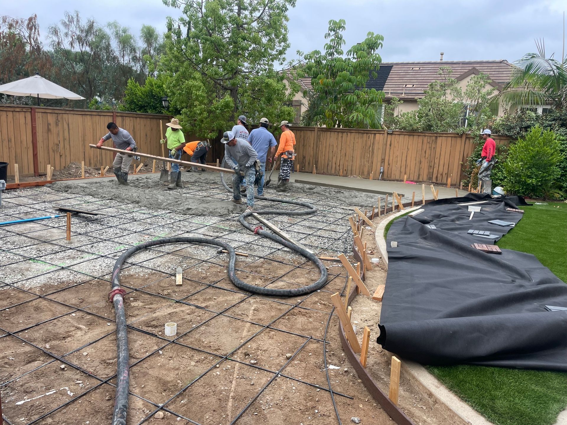 Construction workers pouring concrete on a prepared foundation with rebar. Outdoor setting with a fence and grass.