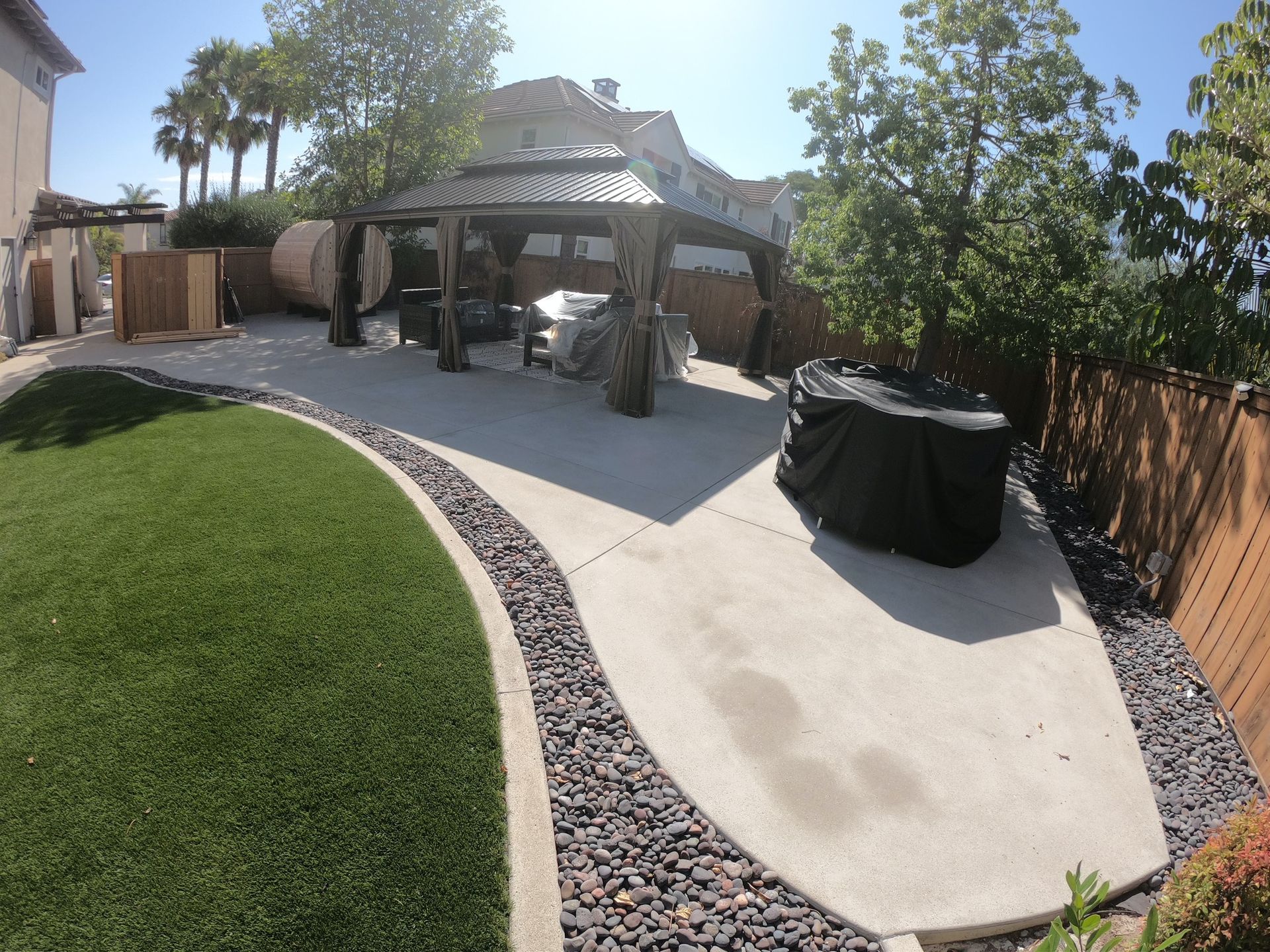 Backyard with gazebo over patio, artificial grass, stone border, trees and covered grill.