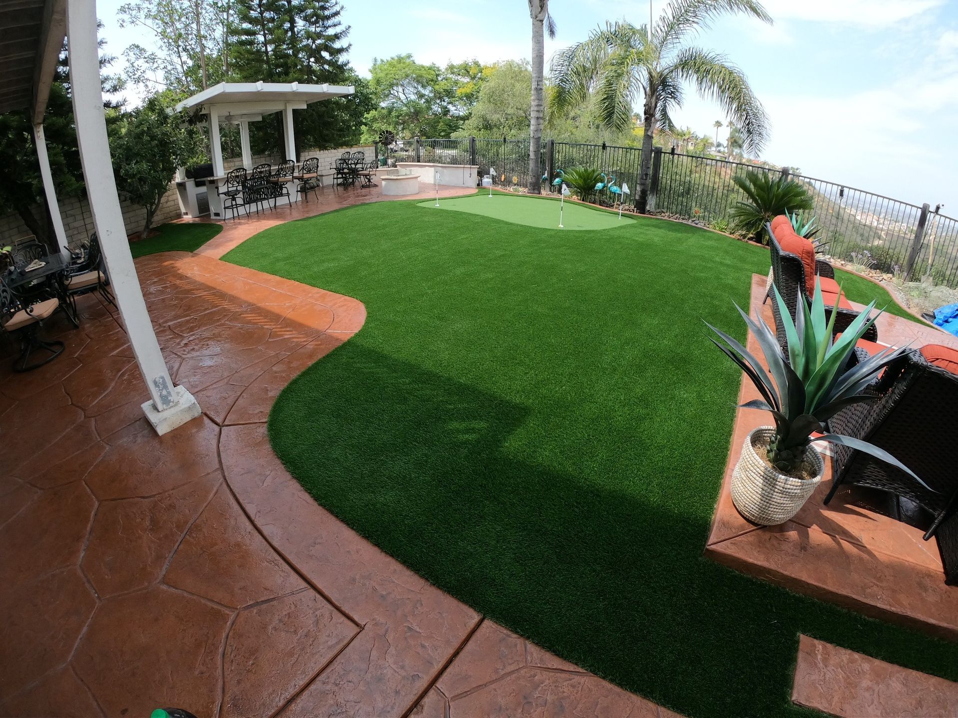 Backyard with artificial turf, brown patio, and gazebo. Green lawn, trees, and sunny sky.