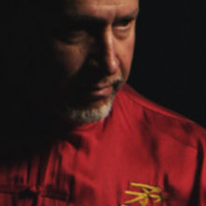 Man in red shirt, looking down, with a partially visible gray beard. Dark background.