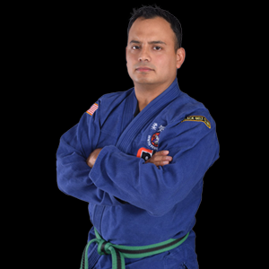Man in blue martial arts uniform with crossed arms, green belt, black background.
