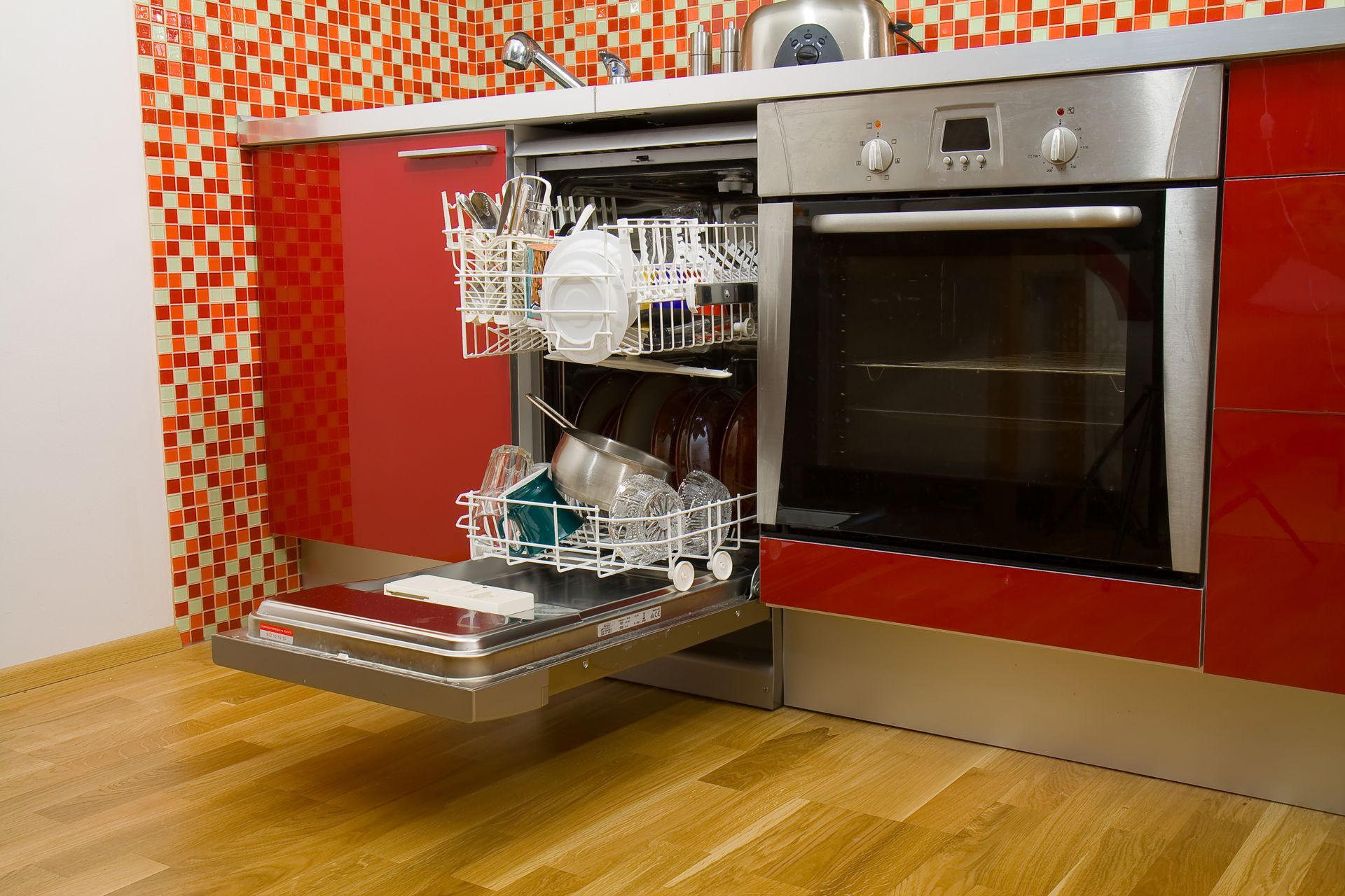 Dishwasher open, filled with dishes, next to oven in a red kitchen with mosaic backsplash.