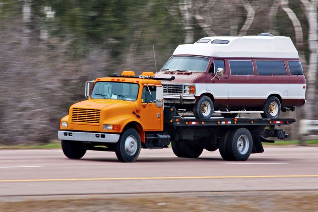 Orange flatbed tow truck carrying a white-and-maroon van on a road.