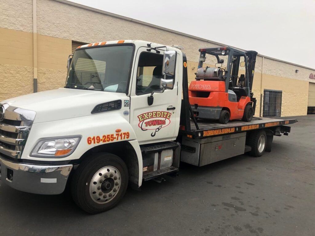 Tow truck carrying an orange forklift parked beside a beige warehouse.
